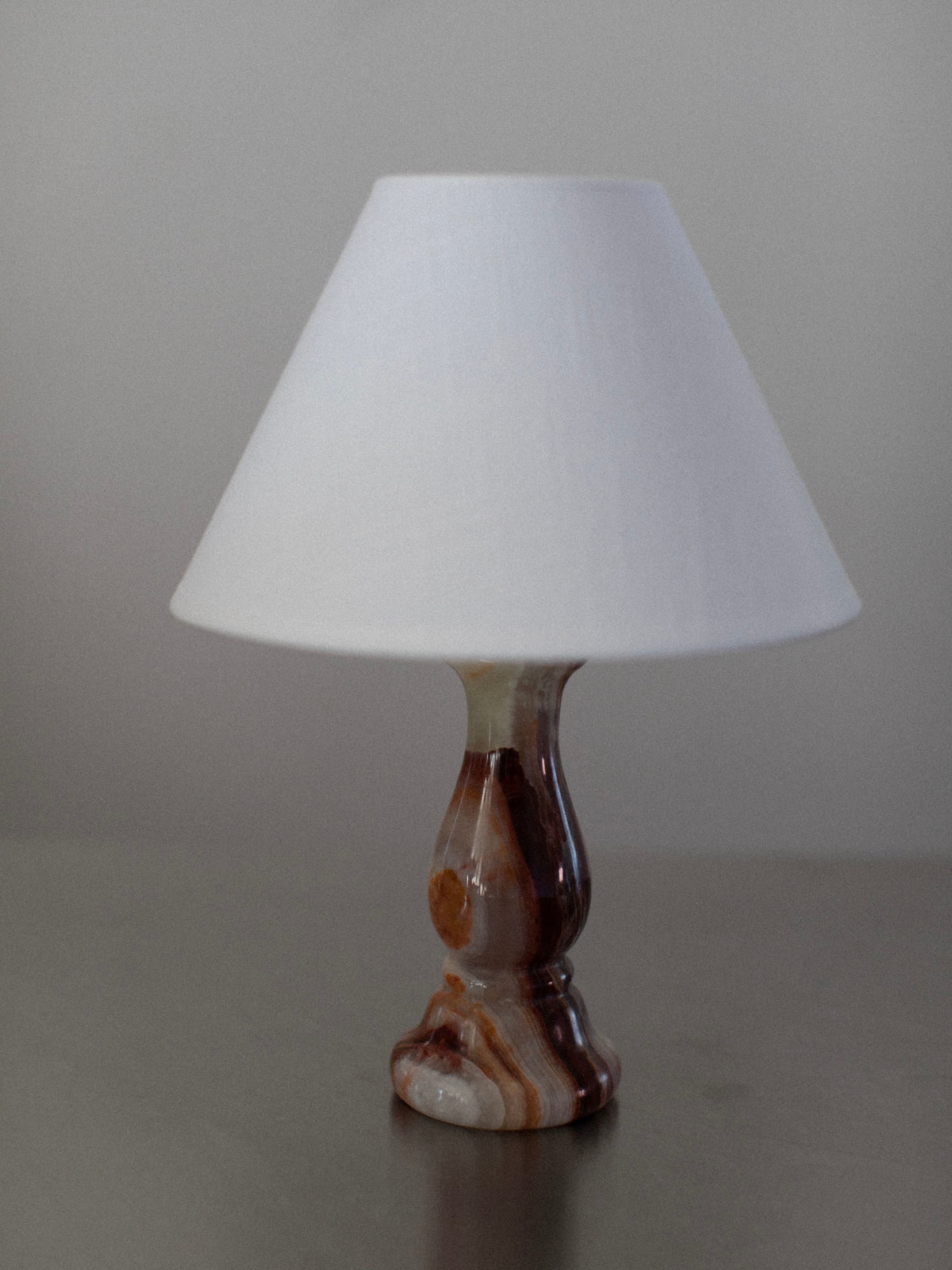 A small table lamp from the Marble Table Lamps Duo by Out For Lunch, featuring a white conical lampshade and a decorative base with shades of brown and cream marble, is placed on a smooth, neutral-colored surface against a plain gray background.