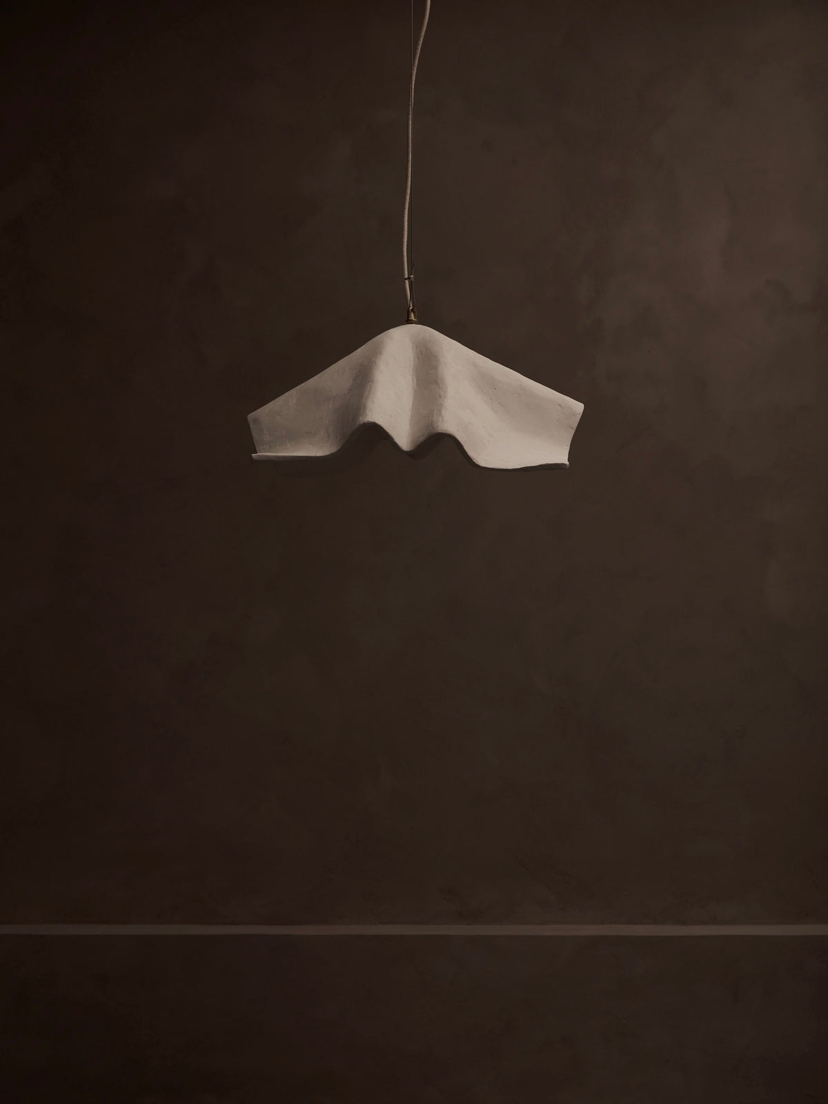 A minimalist image of the 'La Fleur' Pendant Light by Studio Tristan Kallas showcases its unique wavy design with floral silhouettes, hanging against a dark, textured background. Its dimmable lighting creates a striking contrast with the muted surroundings.
