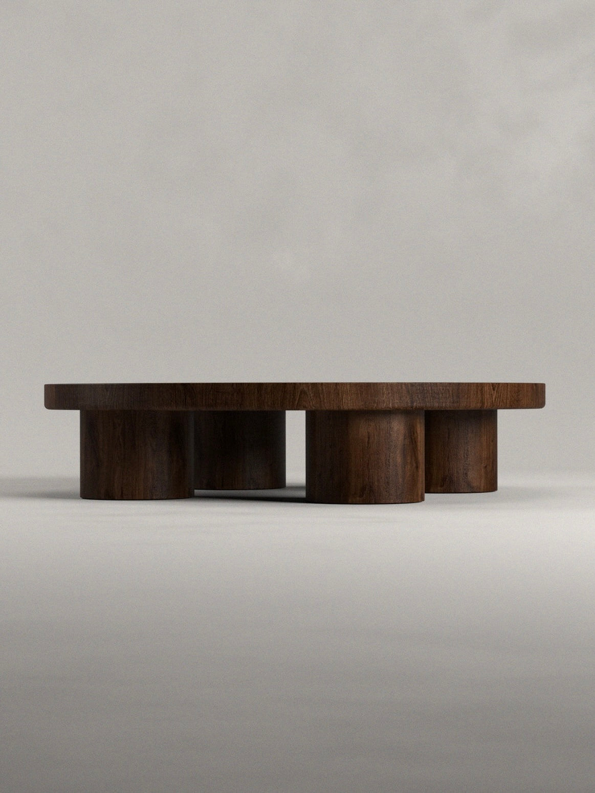The Totem Coffee Table by Siete Studio is a round, dark wooden piece with a thick solid walnut top and cylindrical legs. Its minimalist, sturdy handcrafted design stands out against a plain light gray background.