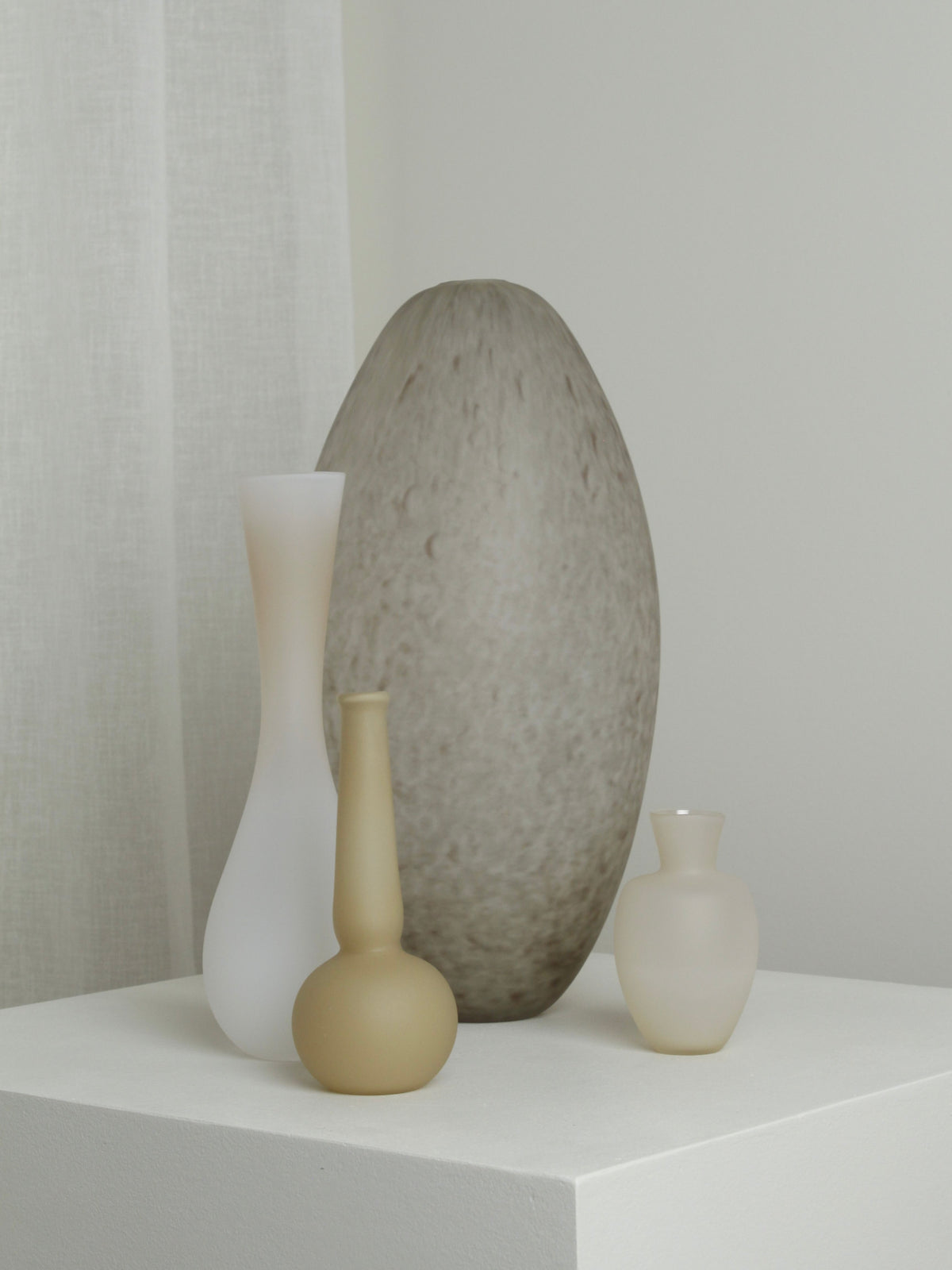 Four minimalist vases of varying sizes and shapes, including the Medium Vase in Mushroom by Verre d'Onge, are displayed on a white pedestal. The vases are made of muted neutral-colored materials, such as translucent frosted glass and ceramic, creating a simple and elegant arrangement against a light background.