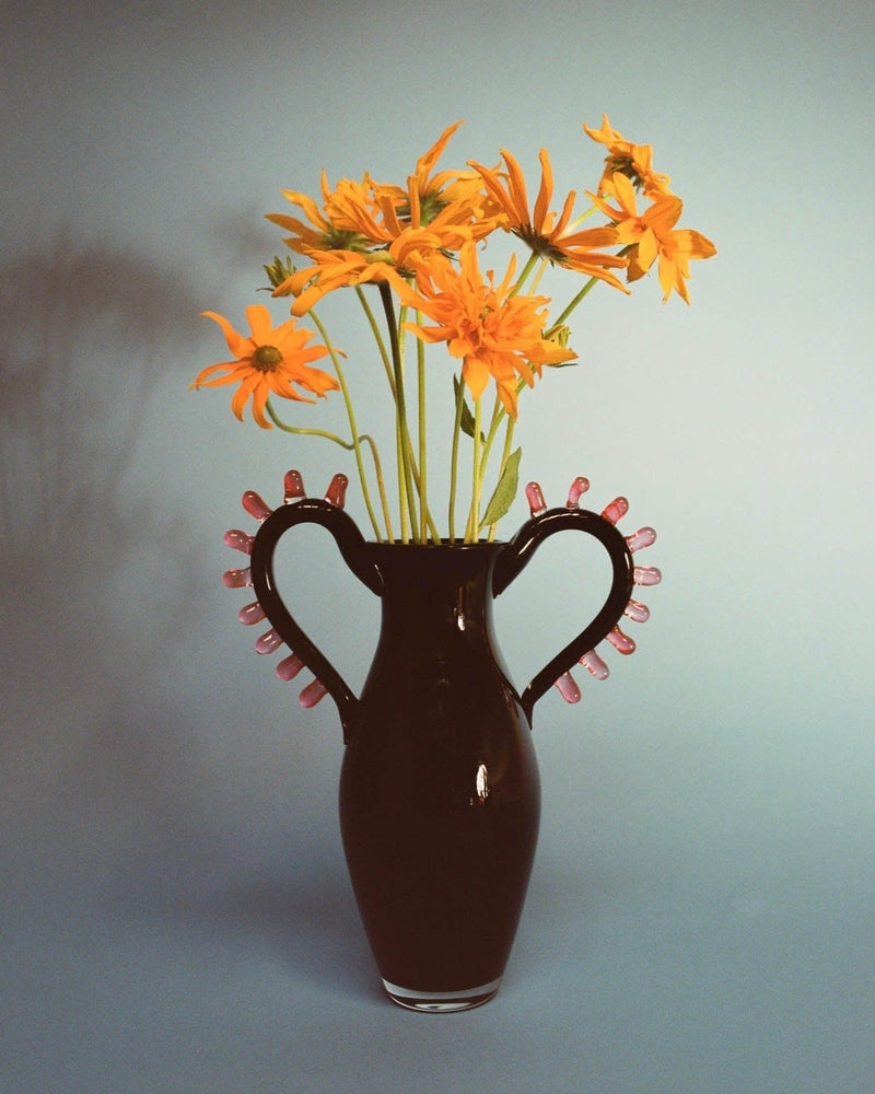 The L'Aurore Vase by Sophie Lou Jacobsen, featuring dual handles and pink accents, holds an artisanal array of vibrant orange flowers. Its soft blue backdrop offers a harmonious contrast, highlighting the minimalist design as flower shadows gracefully move on the background.