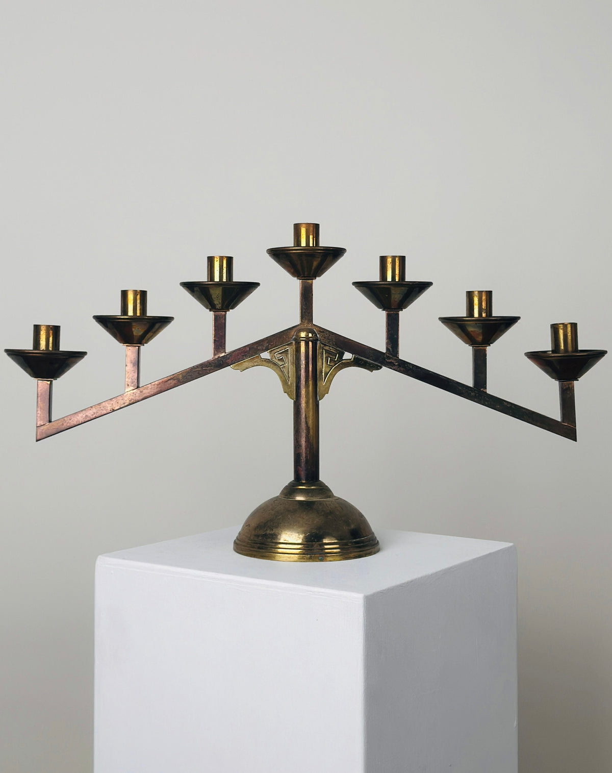 Displayed on a white pedestal against a plain light grey wall, the Modernist Candleholder by Figuresdesign showcases a minimalist and geometric design. This brass candelabra features eight branches along with one central elevated branch, all boasting a beautiful patina that adds character.