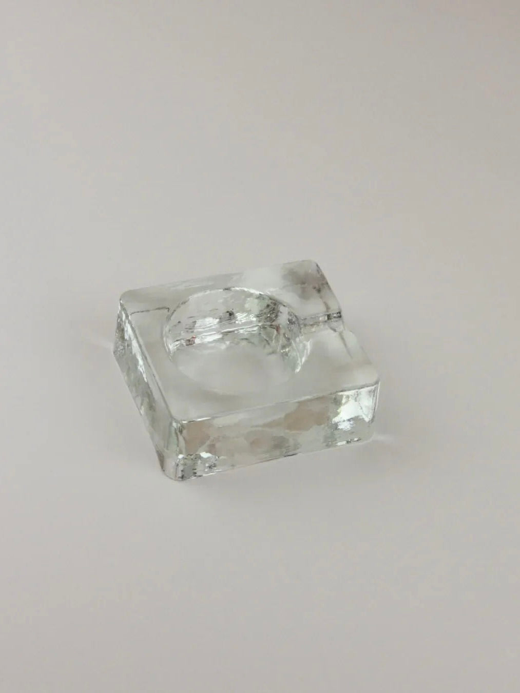 A small, clear glass object with a square base and a circular indentation in the center is shown on a light, neutral background. The Brutalist Ashtray from Boga Avante Shop has facets that reflect light, giving it a sparkling appearance.