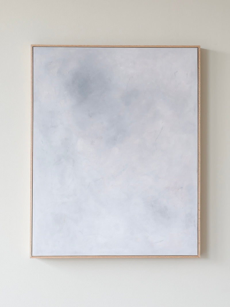 The "Eos" acrylic painting on canvas from A Nesting Place, created in 2024, features a minimalist design with a light-colored surface predominantly showcasing subtle shades of white, gray, and very pale blue. The artwork is elegantly framed in a thin oak frame and is displayed on a light beige wall, exuding a calm and serene aesthetic.