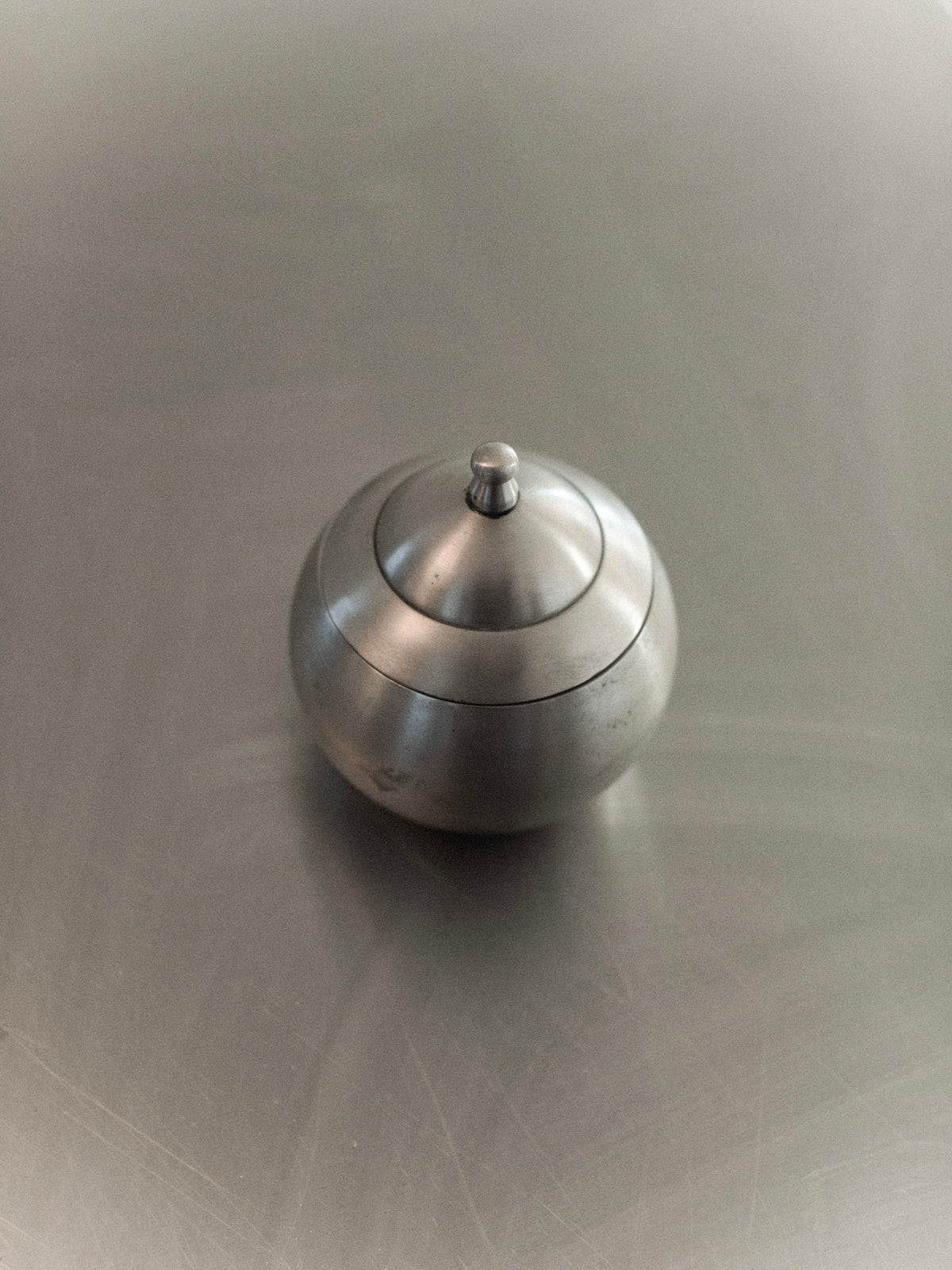 The Steel Bonbonnière from Out For Lunch, a compact metallic sphere with a lid, rests on a sleek reflective surface. Its minimalist and contemporary design radiates industrial elegance, as the gentle lighting casts delicate reflections and shadows on the polished steel.
