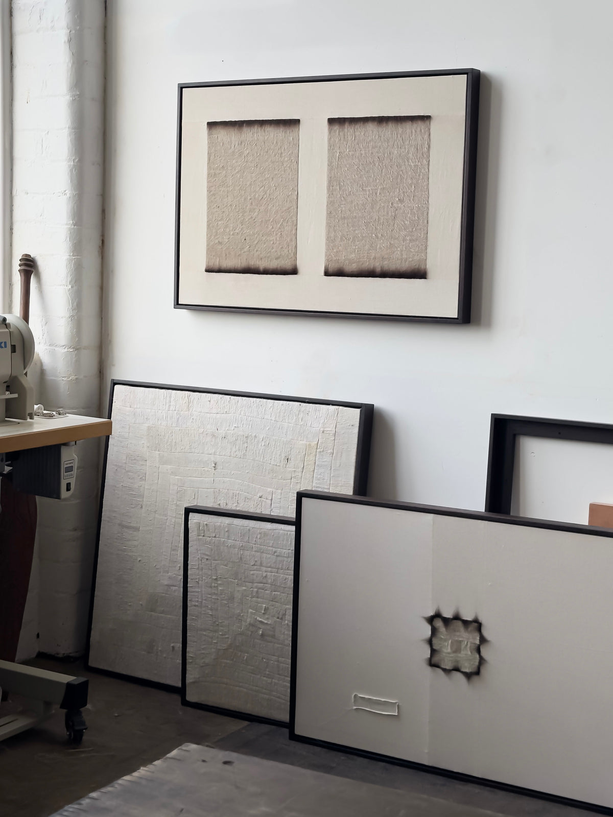 Several framed, textured abstract artworks occupy a corner of an art studio. On the wall hangs "Twin," 2025 by Bec Kirby, highlighting two rectangular textures. Other pieces, including burned art on cotton linen, rest against the wall near a sewing machine and table.