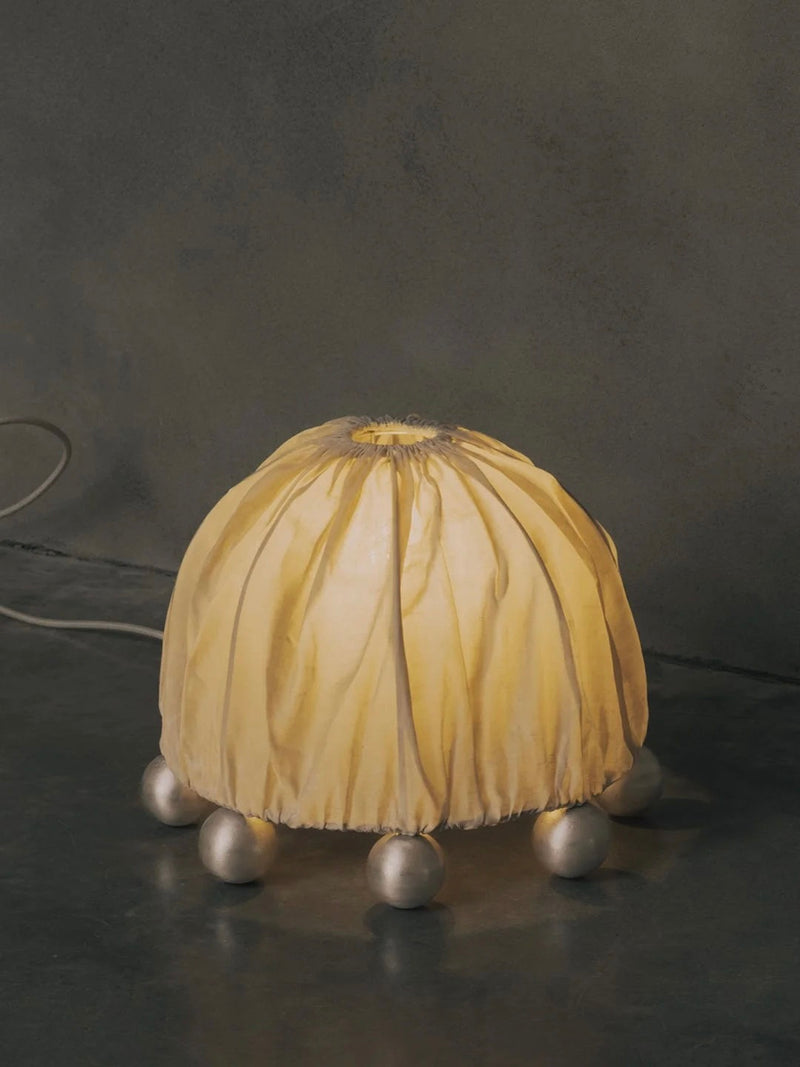 The "ALTER" Table Lamp by Kaytar features a fabric-covered dome emitting a warm glow, supported by spherical feet. With its sculptural design on a dark reflective surface against a muted background, it exudes an artistic charm reminiscent of alchemy symbols.