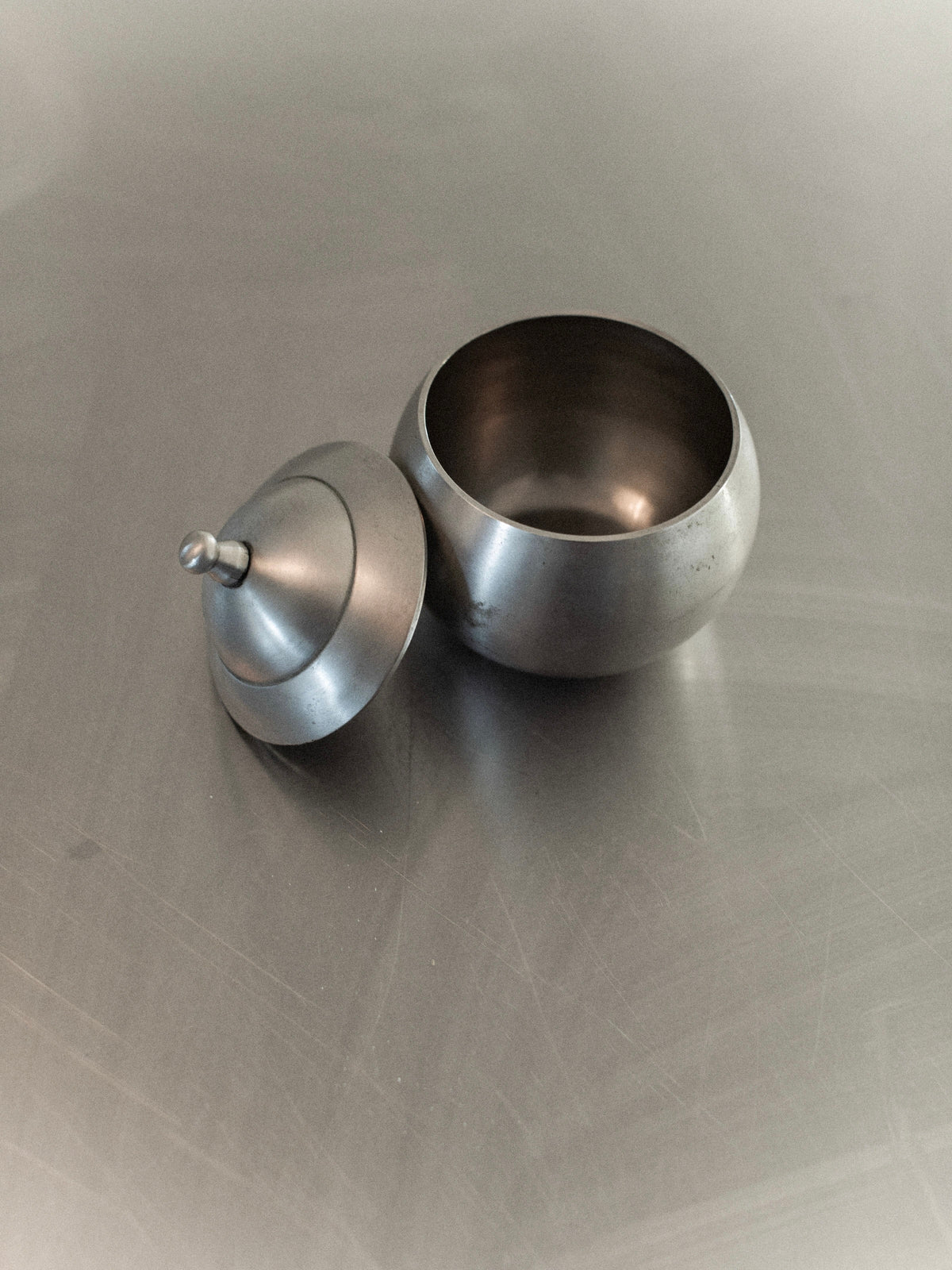 An Out For Lunch Steel Bonbonnière crafted from polished stainless steel rests on a sleek, reflective surface. The round, open container exposes its hollow interior, while the lid with a delicate handle is positioned next to it, radiating mid-century elegance.