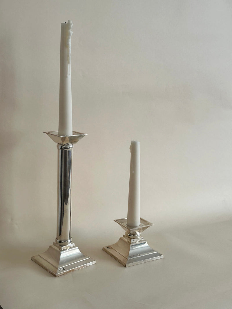 A tall, partially melted white candle stands in an Aesthete Label vintage candle holder on the left, while a shorter, more melted white candle sits in a similar silver German Candle Holders Pair by Aesthete Label on the right; both boast classical column style and are set against a plain, light background.