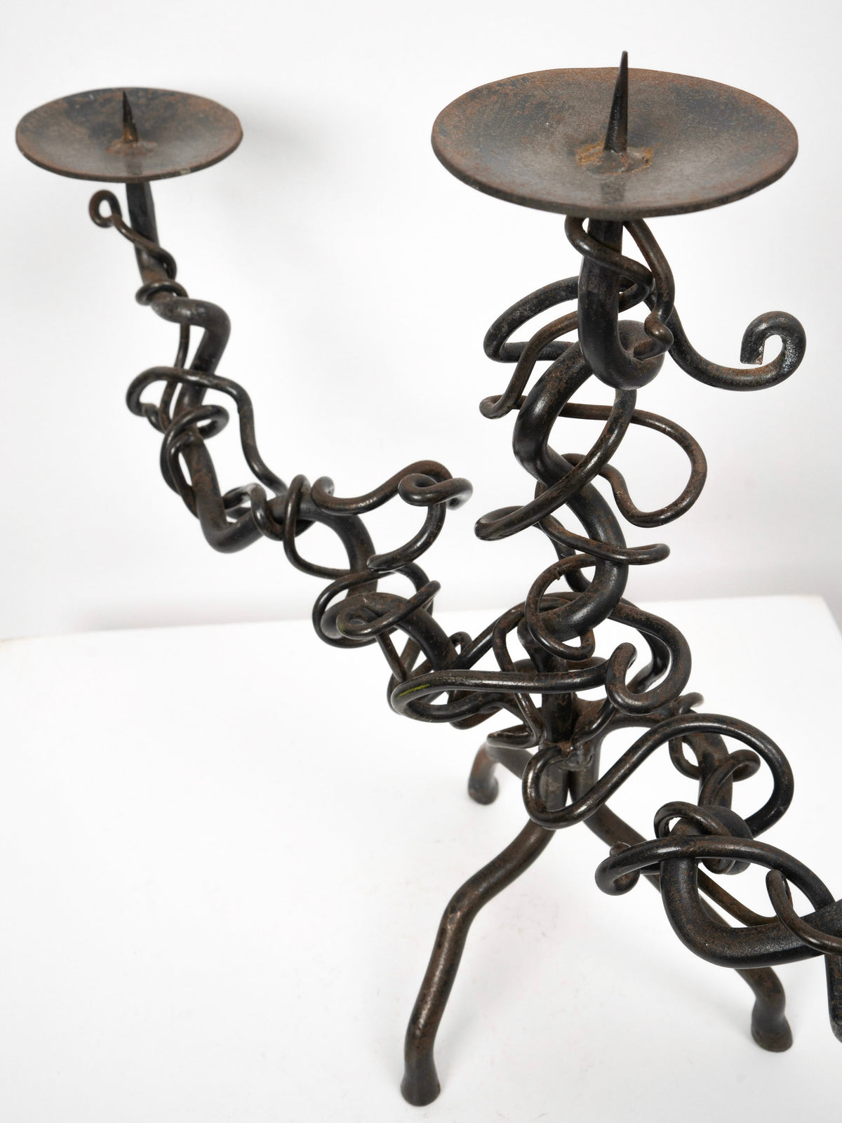 The Brutalist Iron Candelabra by Tom Bogle features two arms with twisted iron rods, flat circular plates with central spikes for candles, and a sturdy three-legged base.