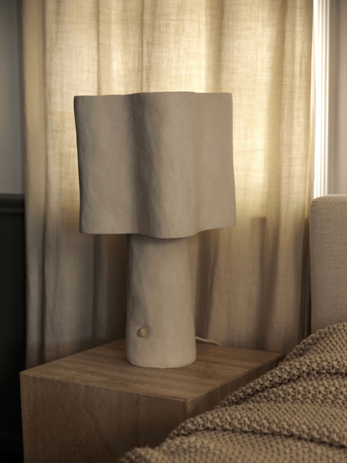 A sculptural Studio Tristan Kallas Freeform Table Lamp with a wavy shade rests on a wooden nightstand. Sheer beige curtains in the background filter soft light, and part of a knitted blanket is visible on the bed to the right.