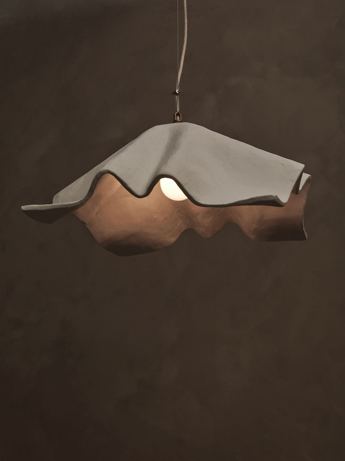 The 'La Fleur' Pendant Light by Studio Tristan Kallas hangs from the ceiling, boasting an irregular wavy white shade. It emits a warm glow against dark settings and offers dimmable lighting for customizable ambiance.