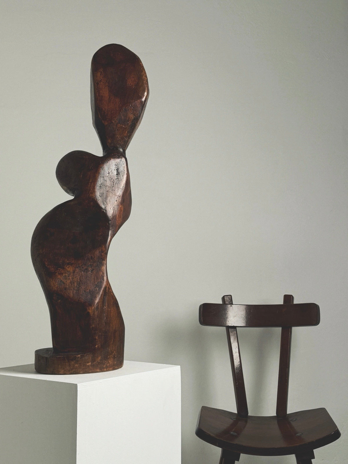 The Large Anthropomorphic Wood Sculpture by Figuresdesign stands elegantly on a white pedestal, beside a simple wooden chair against a plain gray background. Its dark wood features smooth, rounded shapes and an elongated form, reminiscent of a woman's silhouette.