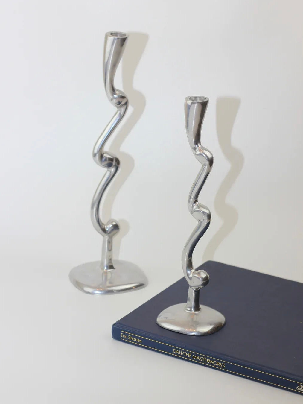 A pair of Boga Avante Shop aluminum candleholders, featuring a sculptural and wavy design, elegantly rests on the hardcover book "Dali: The Masterworks" by Eric Shanes. These pieces contribute a modern flair against the plain white background.