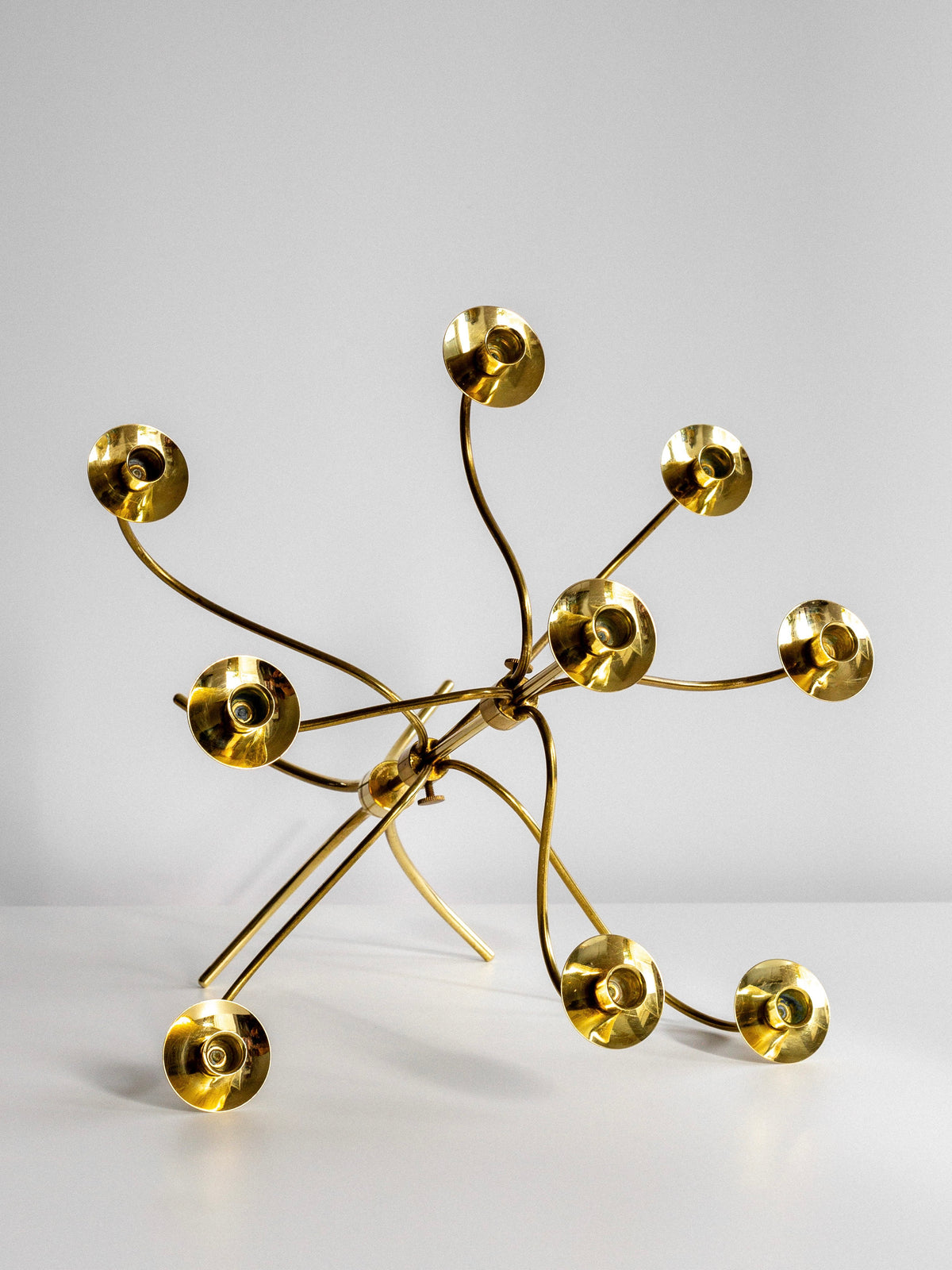 A Large Mid-Century Brass Candelabra by Spigel, reminiscent of a 1950s Josef Frank design, stands on a solid white surface against a plain gray background. The abstract golden metal sculpture features multiple curved stems, each ending in circular, disc-like shapes. The stems are arranged in a radial pattern, creating a sense of dynamic movement and capturing the essence of Svenskt Tenn design.