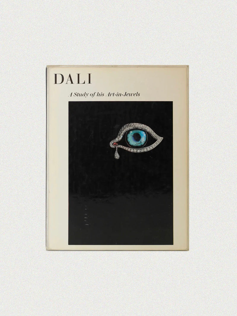 The book cover for "Dalí: A Study of his Art-in-Jewels" by Boga Avante Shop features a surreal eye-shaped jewelry design with a blue iris and teardrop on a black background, celebrating Dalí’s artistic legacy. The title is set in the top left corner.