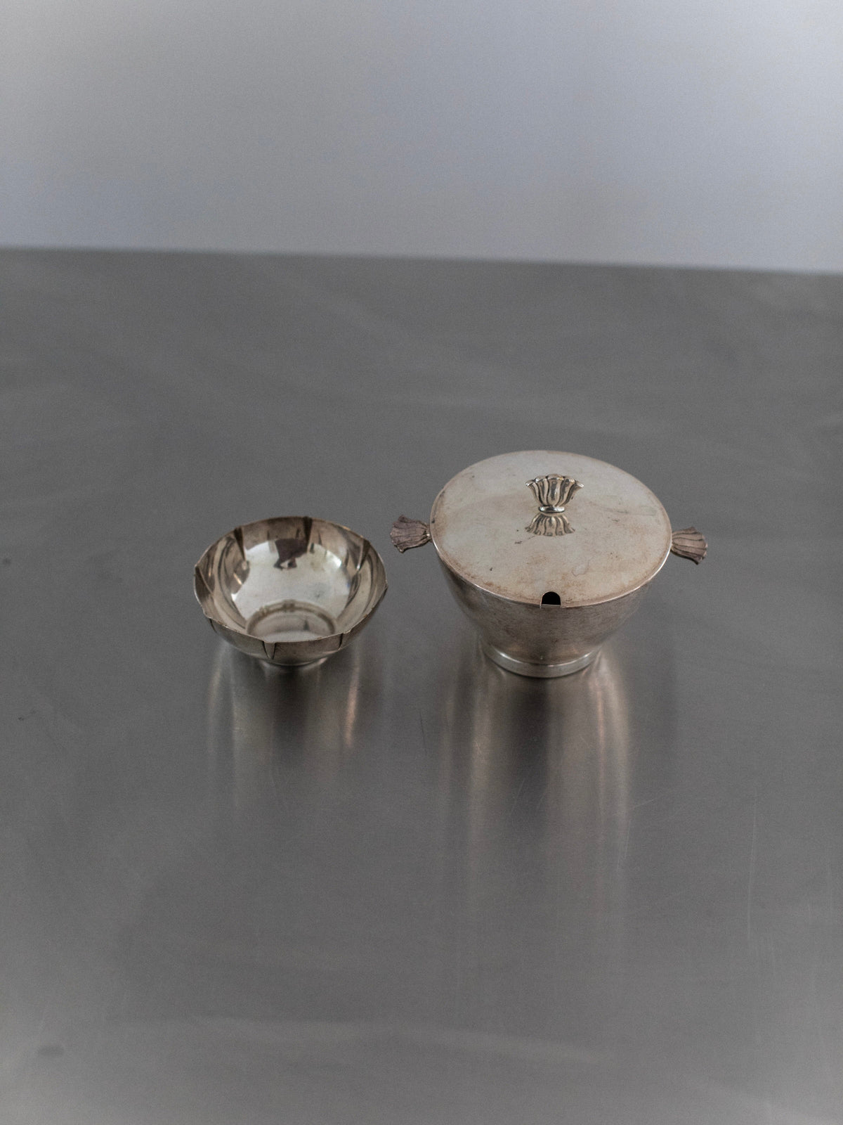 A small silver bowl and a larger Out For Lunch Art Deco-inspired container with a lid rest on a smooth gray surface. The container's lid showcases a decorative element on top and sleek handles on the sides, echoing the style of vintage glamour from a bygone era.