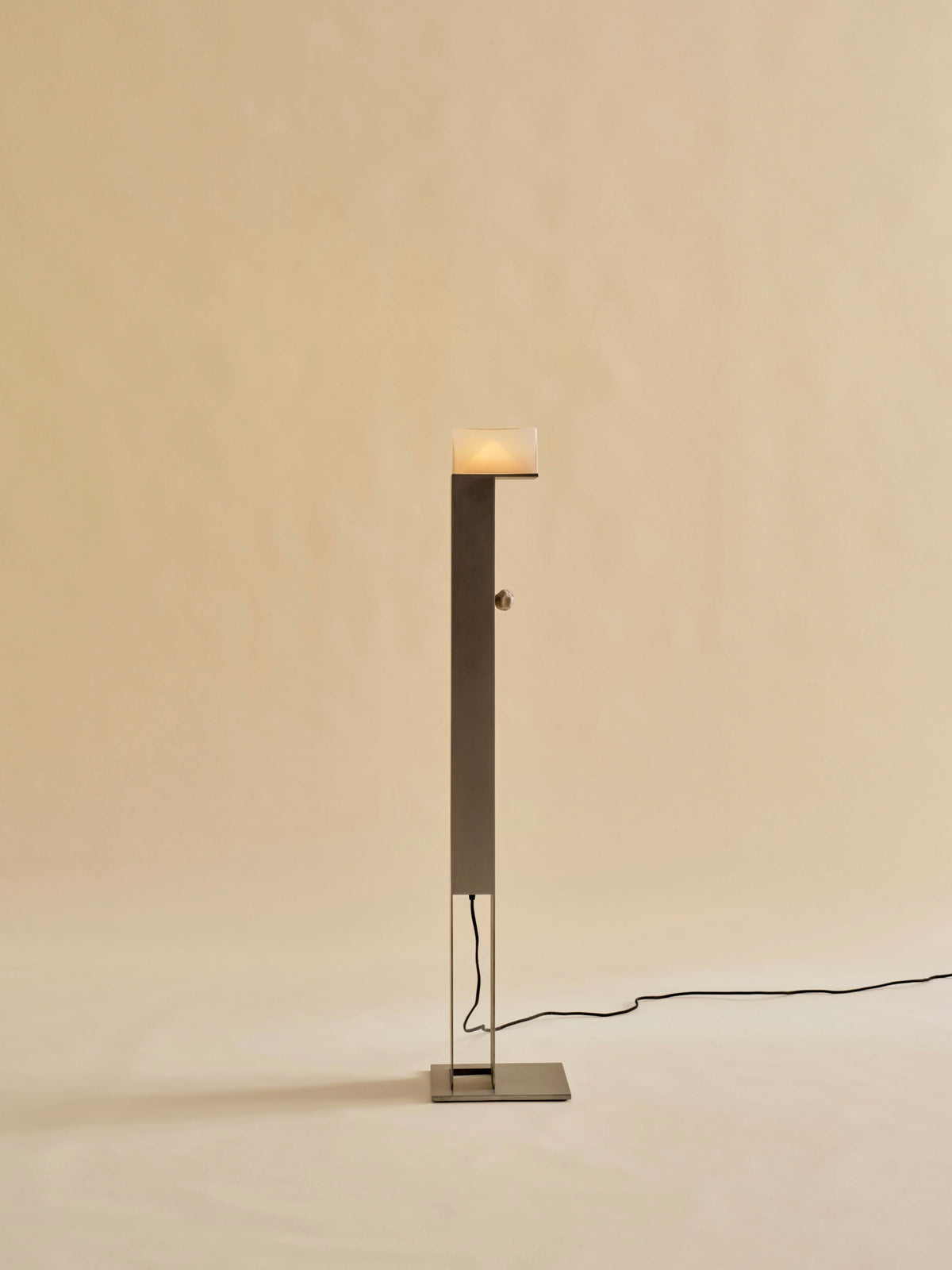 The Perceptions Floor Lamp by Known Work features a minimalist design with a square base, slender pole, and handcrafted cube shade. It casts a soft glow against beige, creating an atmospheric light. A black power cord extends from the base.