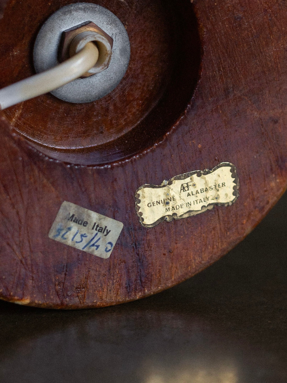 Close-up of the bottom of a vintage Large Italian Alabaster Stone Lamp by Spigel with two stickers. One sticker reads "Genuine Alabaster Made in Italy" in a decorative gold design, and the other, a shorter plain strip, reads "Made Italy 815/0." This neoclassical style piece also features a cord entering through a metal fixture.