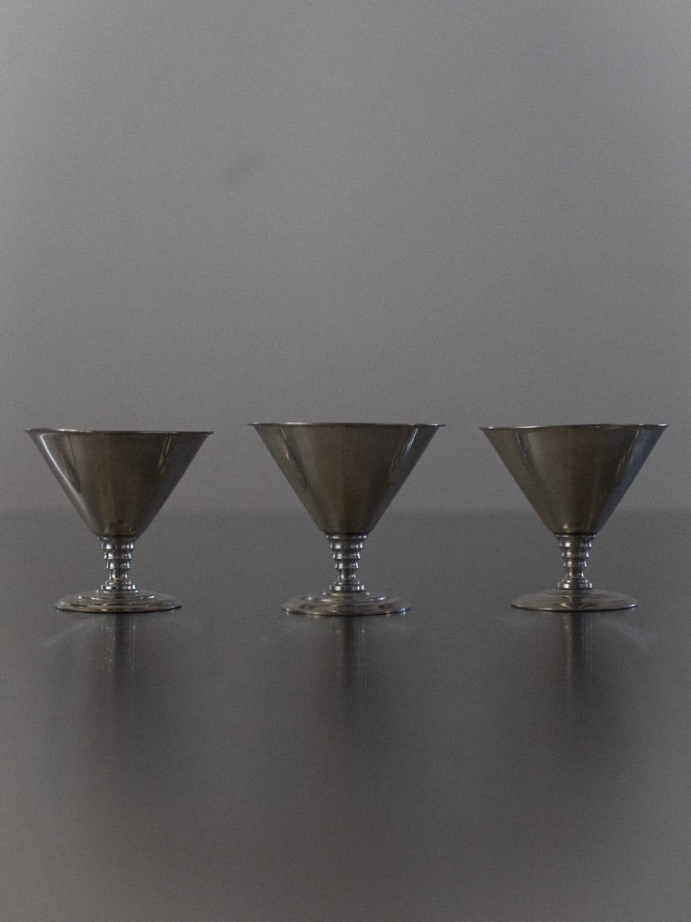 Three identical ice cream cups from the brand "Out For Lunch" are arranged in a row against a plain, gray background. These silver-colored cups, part of the "Six Ice Cream Cups" set, have a conical shape with a wide rim and narrow base, each standing on a small circular pedestal, resembling vintage goblets from Norway.