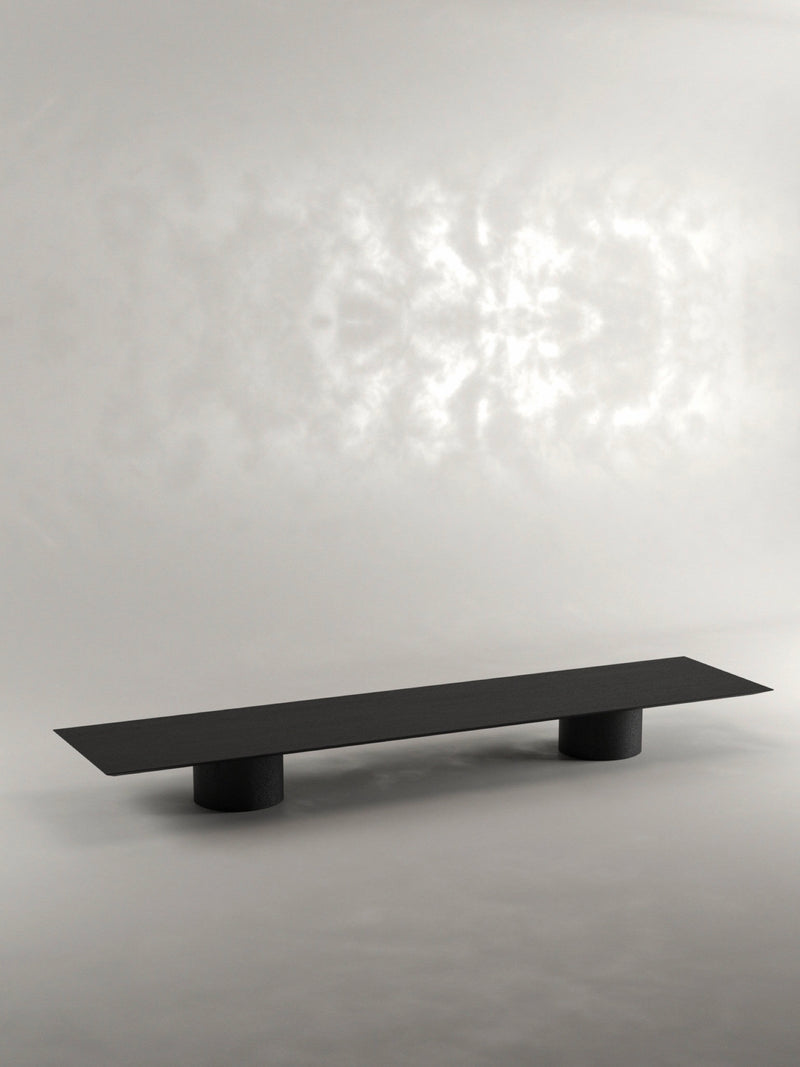 The 'Pier' Coffee Table by Siete Studio is a sleek, minimalist piece handcrafted from black oak with a flat, rectangular surface and two cylindrical legs, set elegantly against a neutral background.