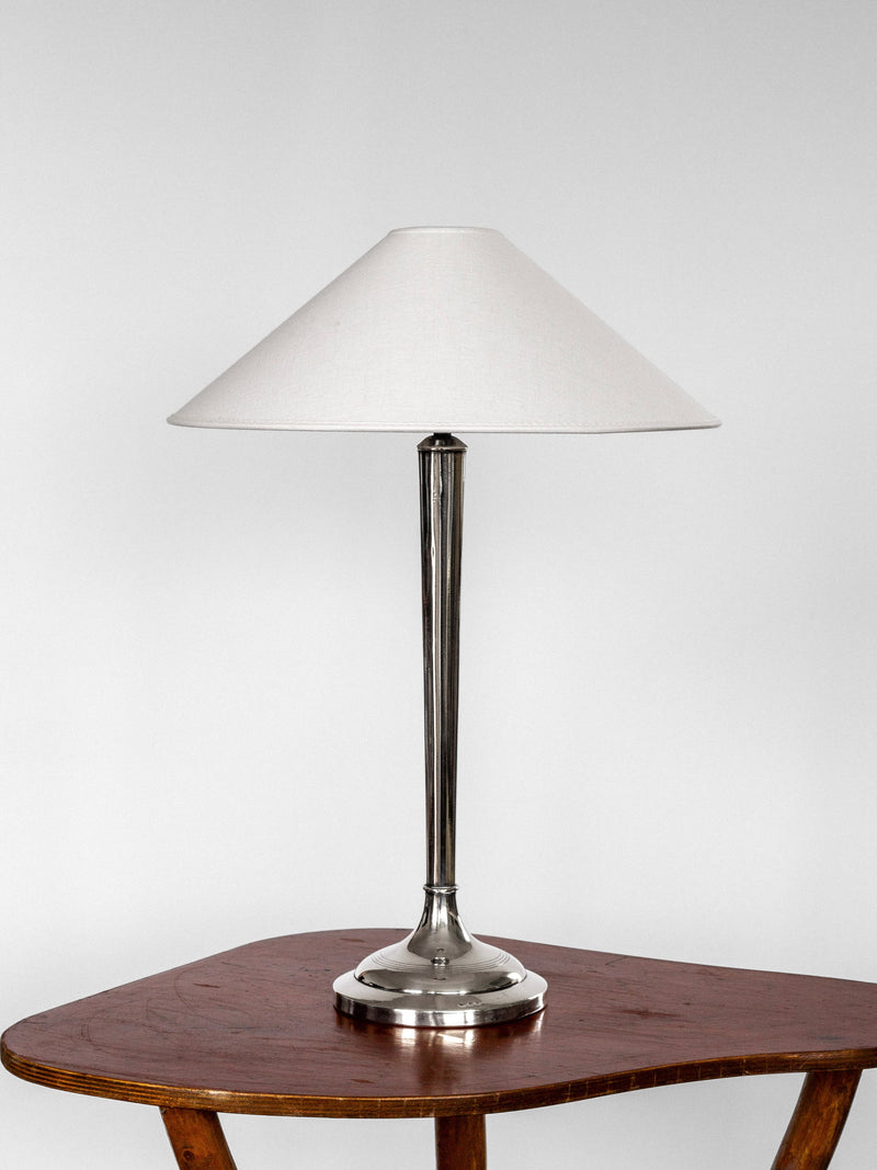 A sleek, modern Silver-Plated Table Lamp from the early 1900s by Spigel boasts a tall, slender metallic base and a wide, white conical lampshade. The lamp is placed on a polished wooden table against a plain, light-colored background.