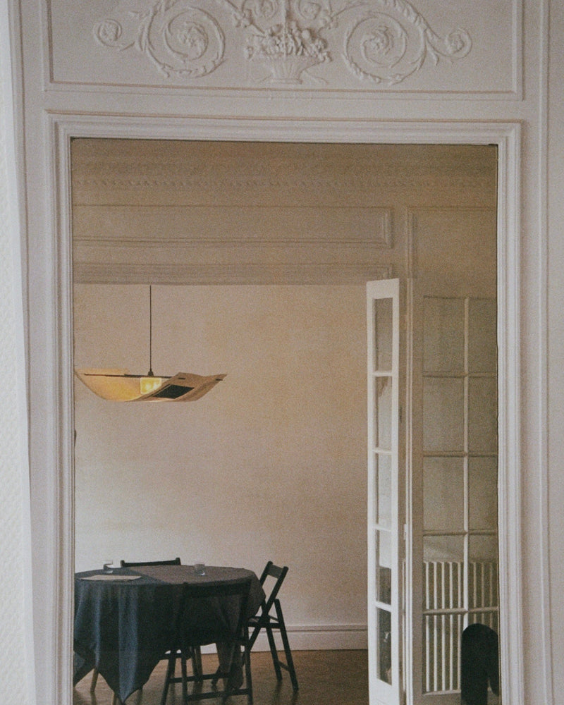 A mirror reflects a dining room featuring a handmade Sail Lamp – Rice Field by TEGET above a round, dark-clothed table. The room includes ornate molding, partially open French doors, minimalist decor, and unique paper collage art.