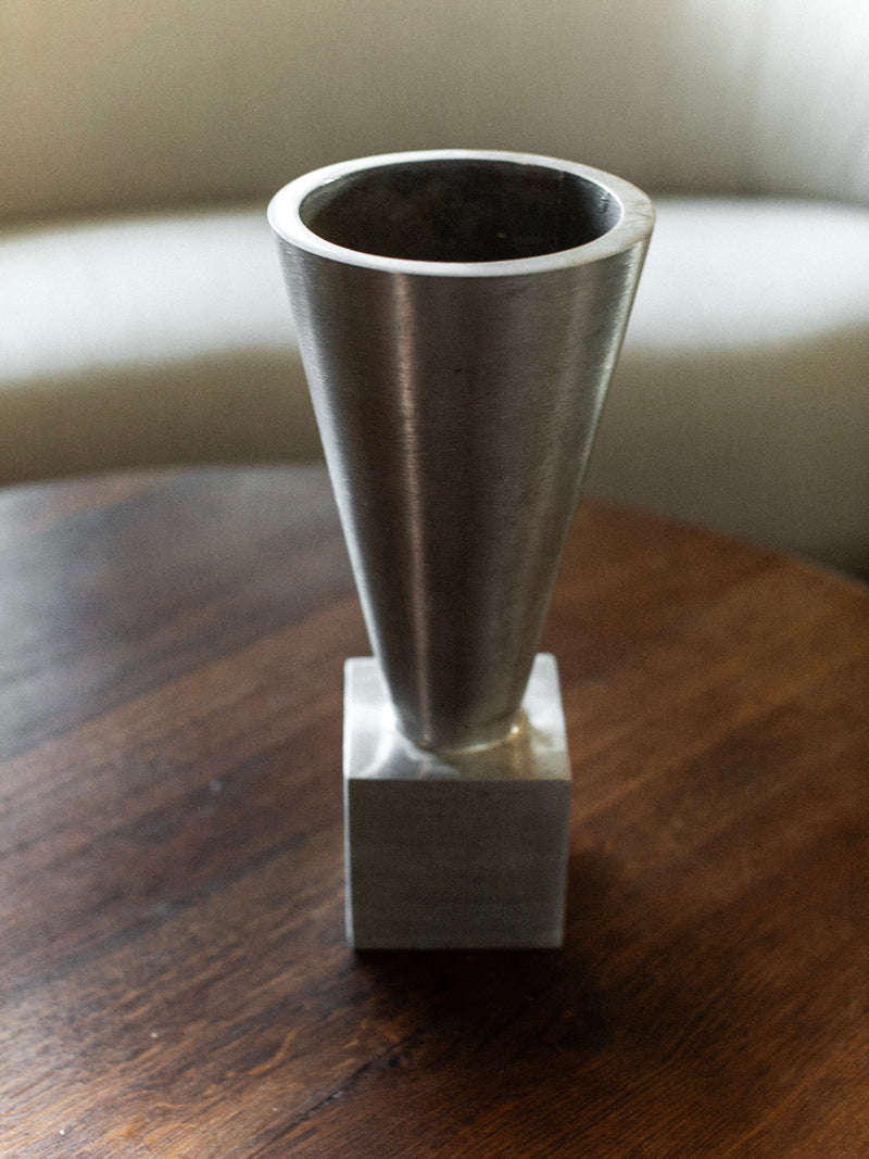 The 70's Aluminum Cast Vase by Out For Lunch, featuring a minimalist cone-shaped form and wide opening in brushed aluminum, sits elegantly on a wooden table. Its modern design provides a striking contrast to the soft, light-colored couch in the background.