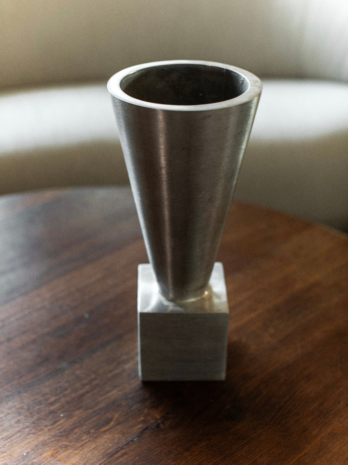 The 70's Aluminum Cast Vase by Out For Lunch, featuring a minimalist cone-shaped form and wide opening in brushed aluminum, sits elegantly on a wooden table. Its modern design provides a striking contrast to the soft, light-colored couch in the background.
