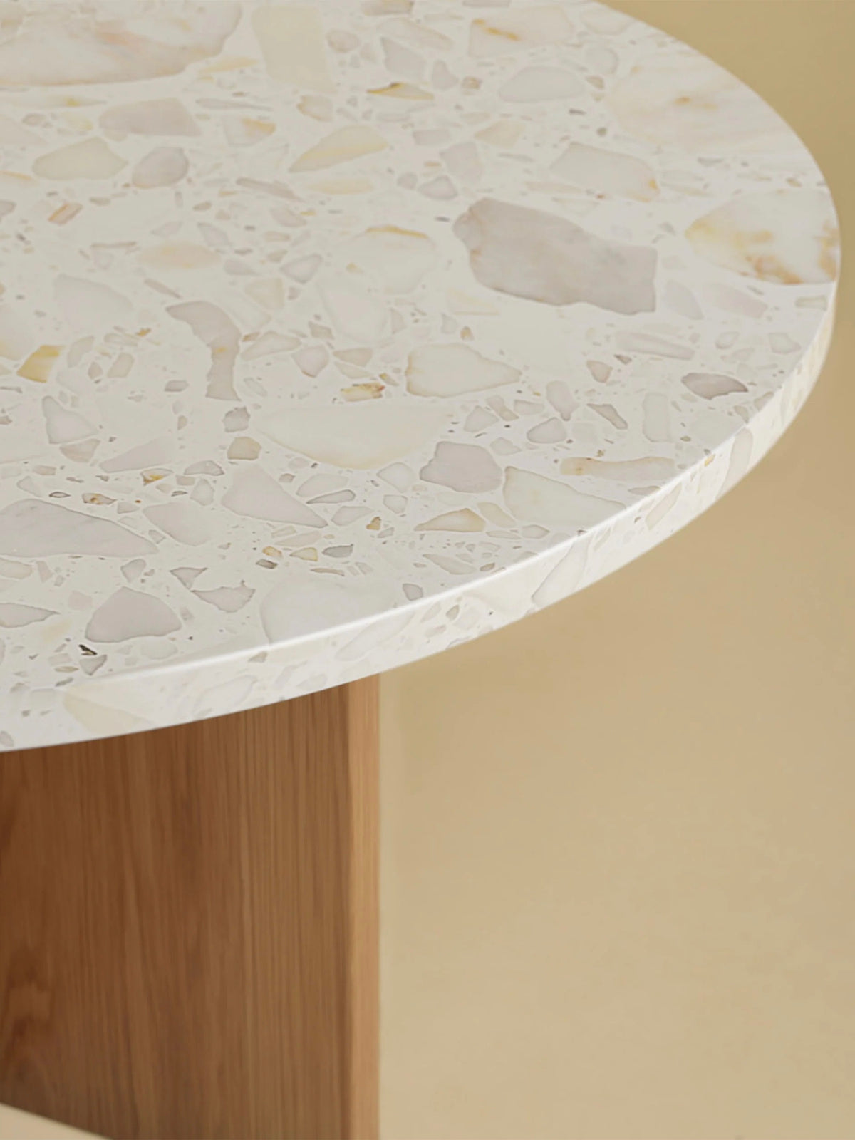 The "Tinian" Coffee Table by Lebanto features a round terrazzo marble top with light gray, beige, and white stone chips. It rests on an oak wood base set against a neutral beige background, highlighting exquisite artisan craftsmanship.