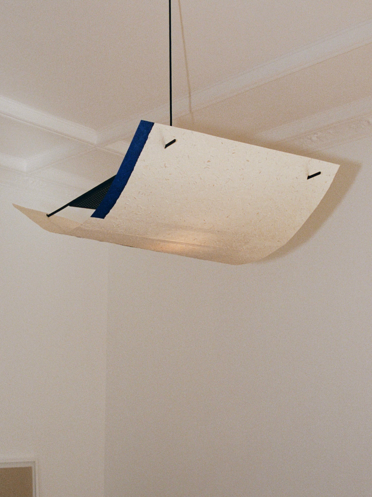 The TEGET Sail Lamp – Rice Field is a modern, minimalist ceiling fixture with a rectangular, slightly curved shade that resembles handmade light-colored material. It hangs artistically by black cords in a neutral-toned room, evoking the layered textures of a paper collage.