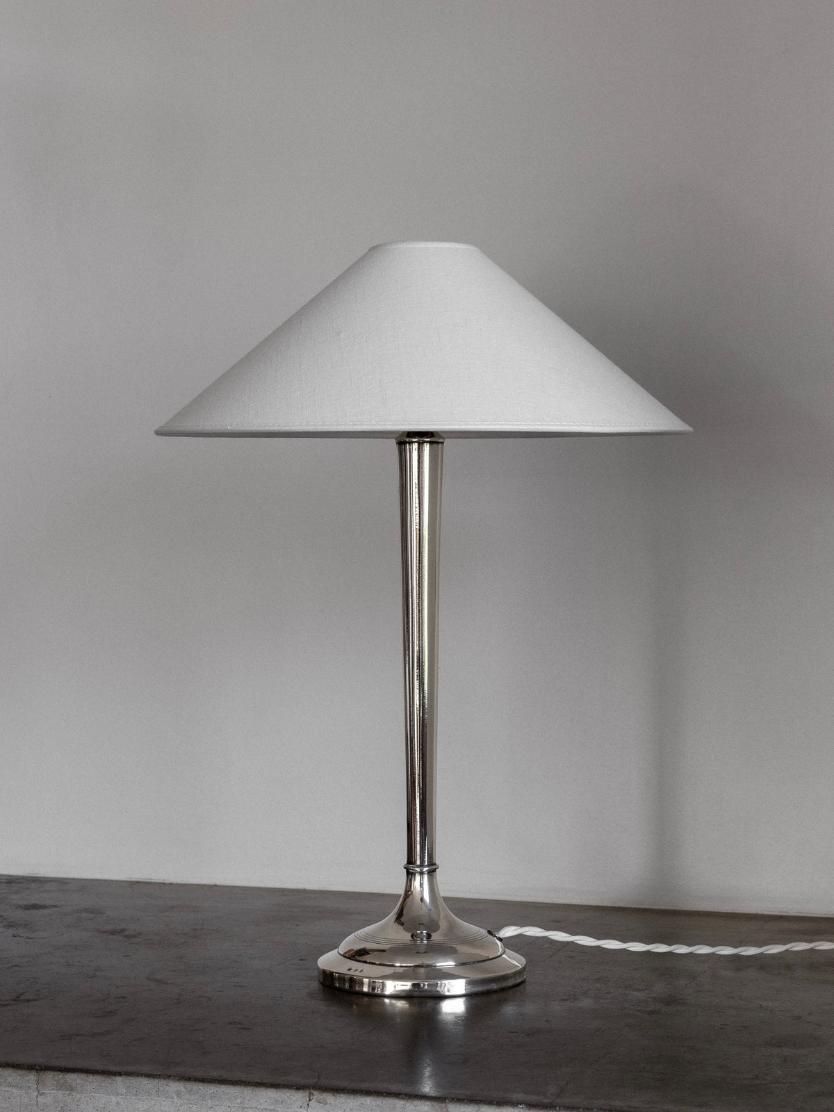 A modern, silver-plated table lamp with neoclassical influences from the early 1900s by Spigel features a sleek metallic base and a conical white lampshade. The lamp stands on a gray stone surface and is turned off, with its power cord trailing behind it against a plain light gray wall.