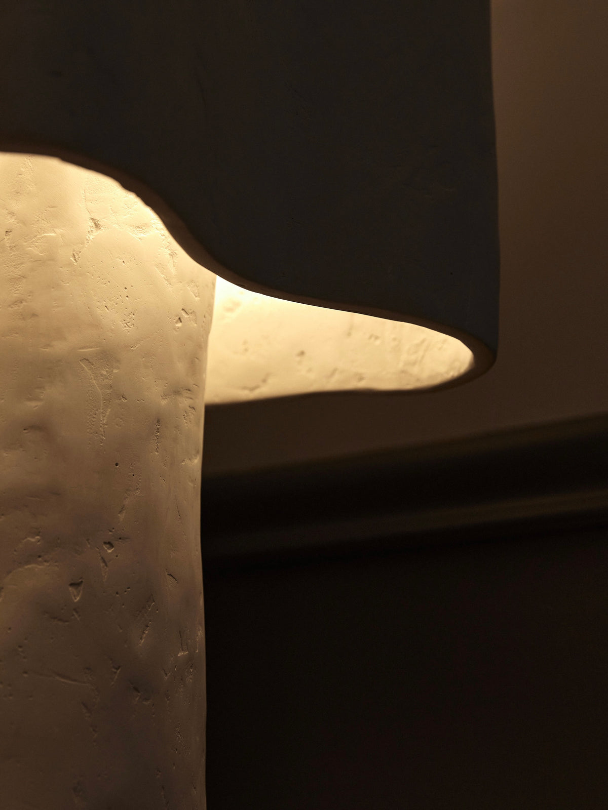 A close-up of the handmade Freeform Table Lamp by Studio Tristan Kallas showcases its textured off-white surface. It emits a warm, soft glow that lights up the surroundings while its shade top creates a gentle shadow contrasting with the light below.