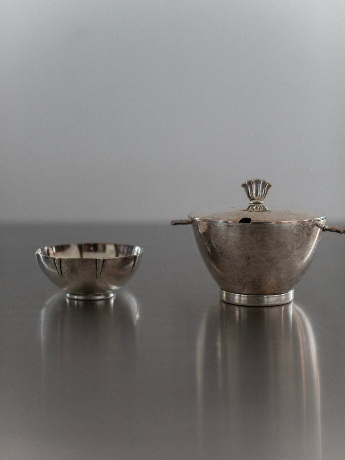 Two Art Deco Bowls from Out For Lunch, one silver and the other a lidded sugar bowl with a decorative handle, rest on a reflective surface. Their polished finish exudes vintage glamour, casting soft reflections that narrate a unique natural story.