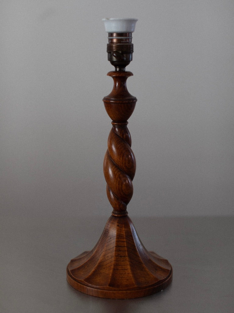 The Early 1900's Table Lamp by Out For Lunch is elegantly showcased against a plain background. This wooden lamp base boasts a polished finish, featuring a fluted bottom and an intricately carved, twisted central column. Topping it off is an empty light bulb socket adorned with a metal band and a white ceramic cap, making it a perfect addition for adding vintage decor to any room.
