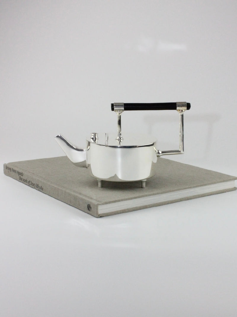 Boga Avante Shop's Christopher Dresser's Tea Pot, a modern silver-plated teapot with a sleek design and an ebonized wood handle, is resting on top of a closed gray hardcover book. The plain white background emphasizes the minimalist aesthetic of the scene, reminiscent of Dr. Christopher Dresser's innovative style.