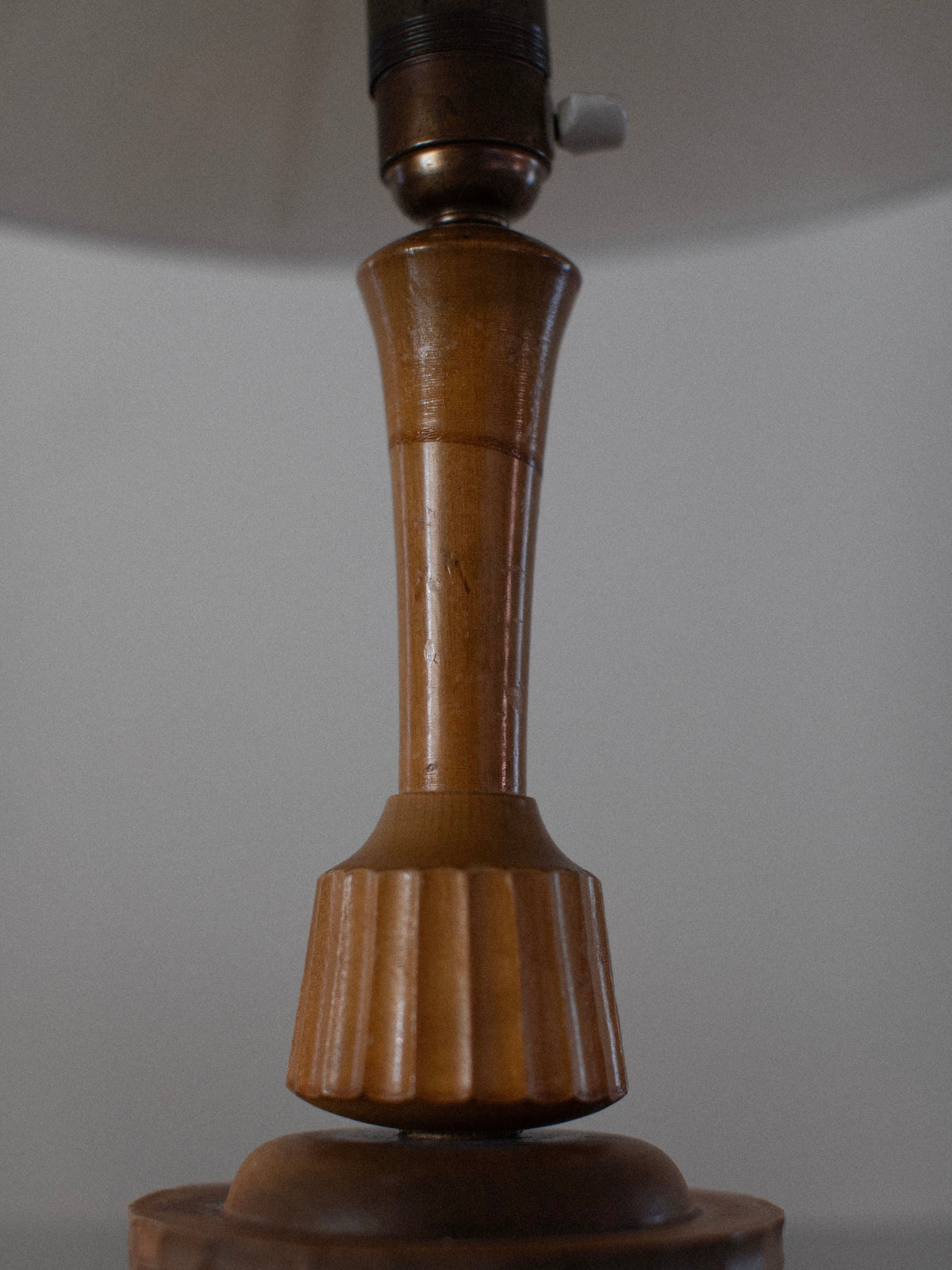 Close-up of the Out For Lunch 1920's Table Lamp, featuring a vintage wooden base with hand-carved details and a tapered column, accented by a fluted design near the bottom. The lamp has a metallic fixture on top with a small toggle switch, all set against a plain, light-colored background.