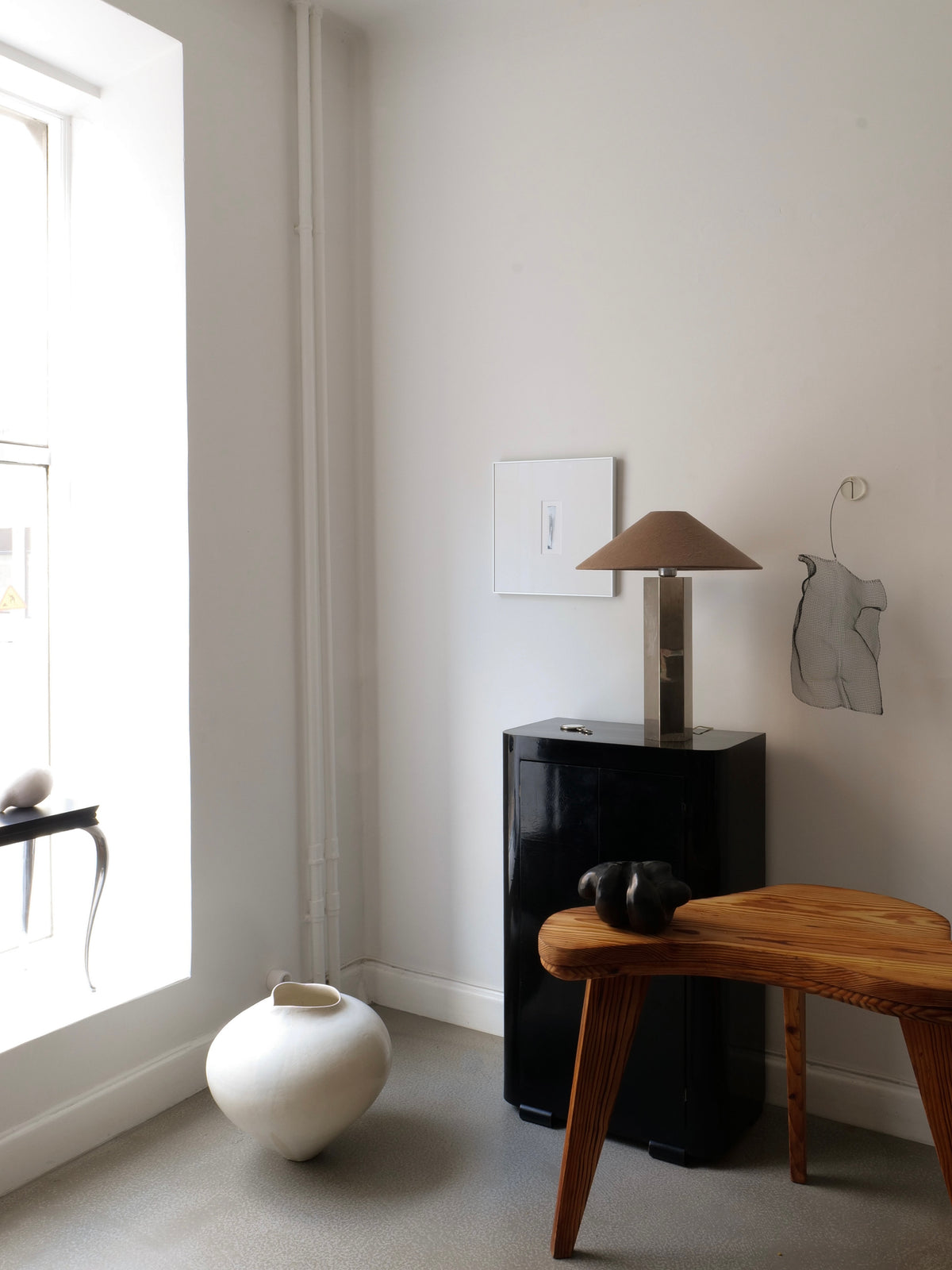 Minimalist room with a white wall, large window, ceramic vase on the floor, wooden table with small black sculpture, black cabinet, Collection apart Grand Hexagonal Lamp, brown shade, and simple abstract wall art.