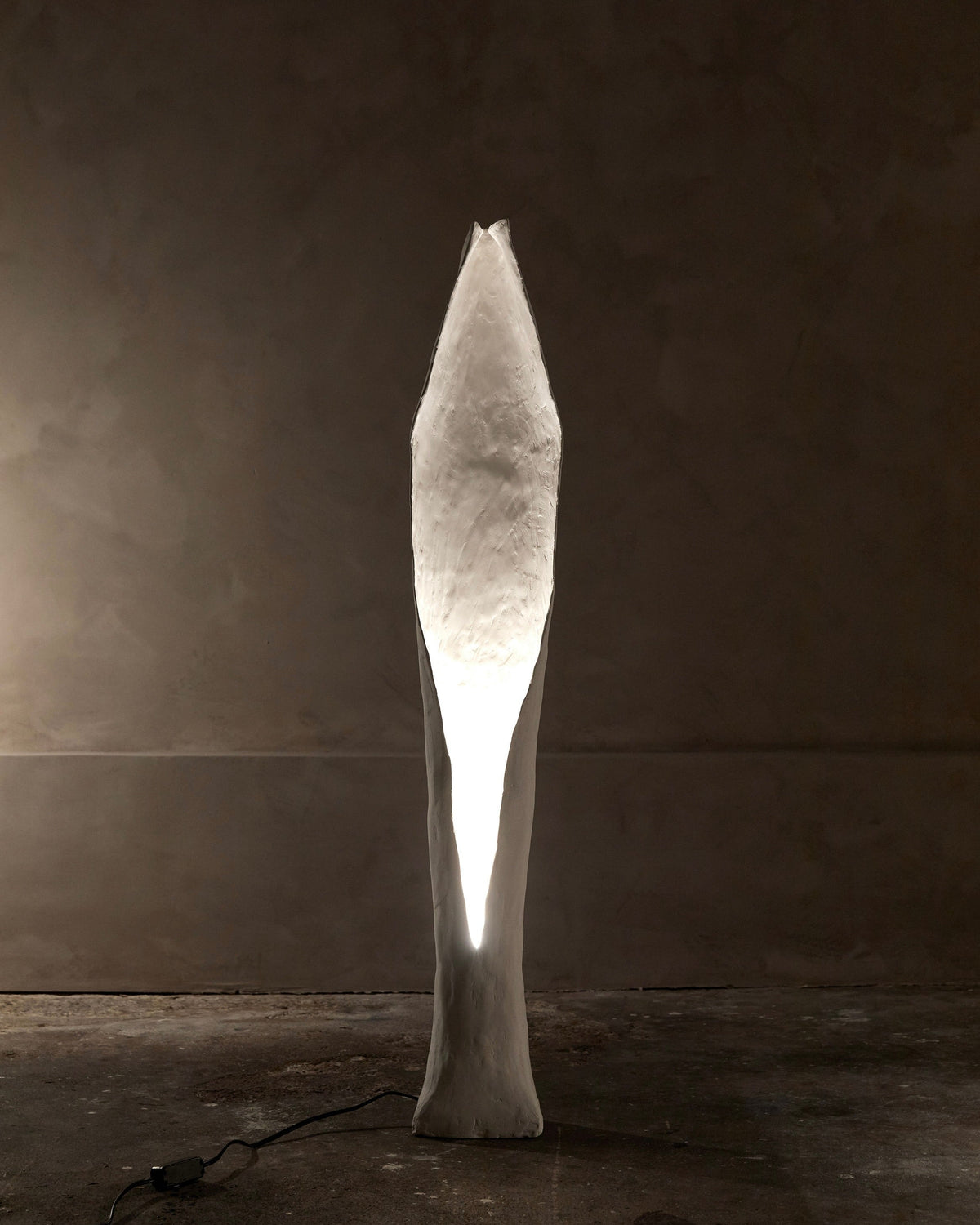A tall, abstract sculpture with a narrow oval-shaped light is reminiscent of the white and textured Alcove Floor Lamp by Studio Tristan Kallas, standing on concrete against a dimly lit, neutral background.