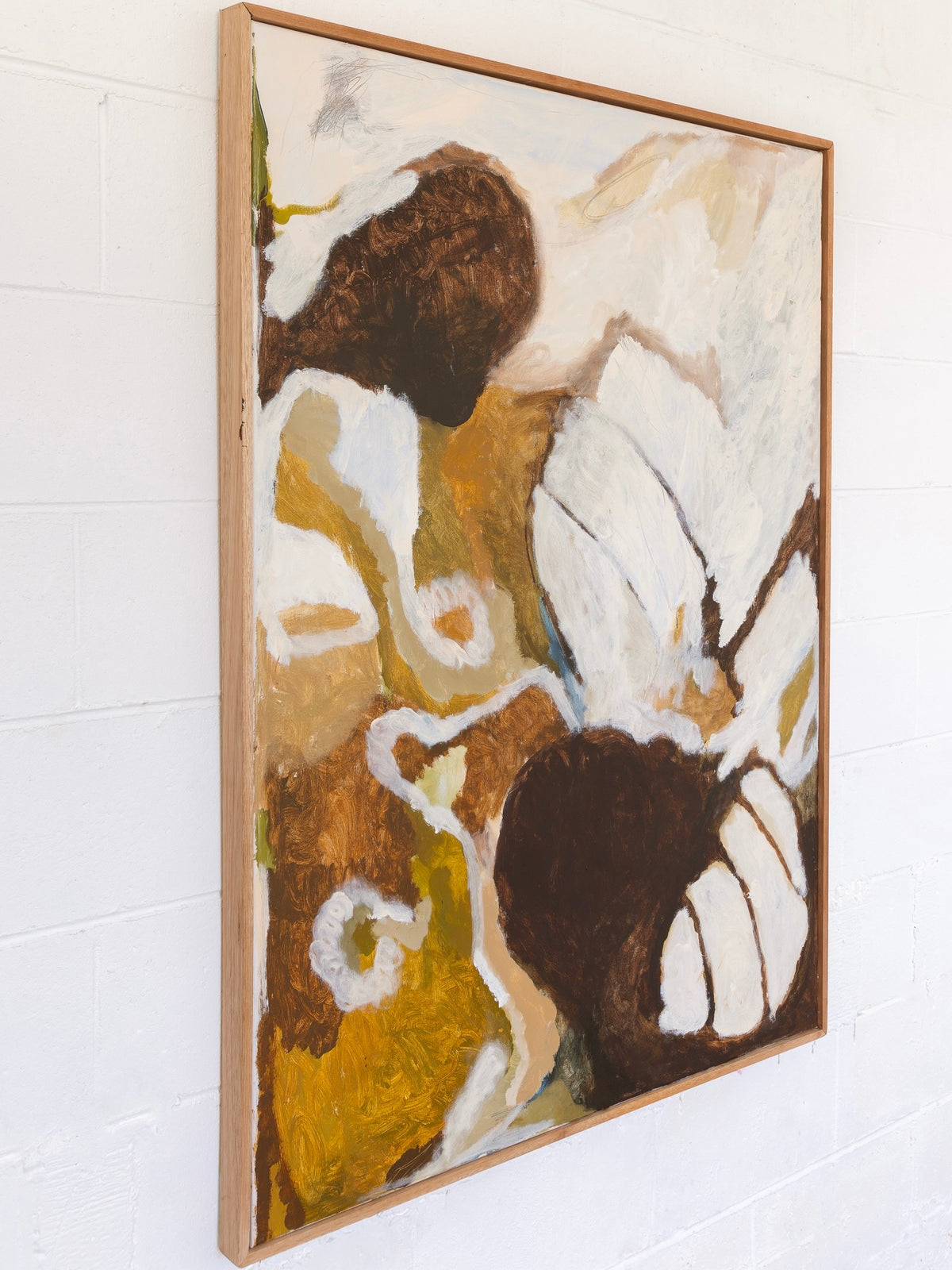 The "I'd Cross The Earth To You - Painting" by Natasha France showcases an abstract design in an earthy color palette of browns, whites, and yellows. The canvas features fluid, swirling shapes and textures that impart a sense of movement and intimate emotion. This artwork is elegantly framed in a simple wooden frame and mounted on a white wall.