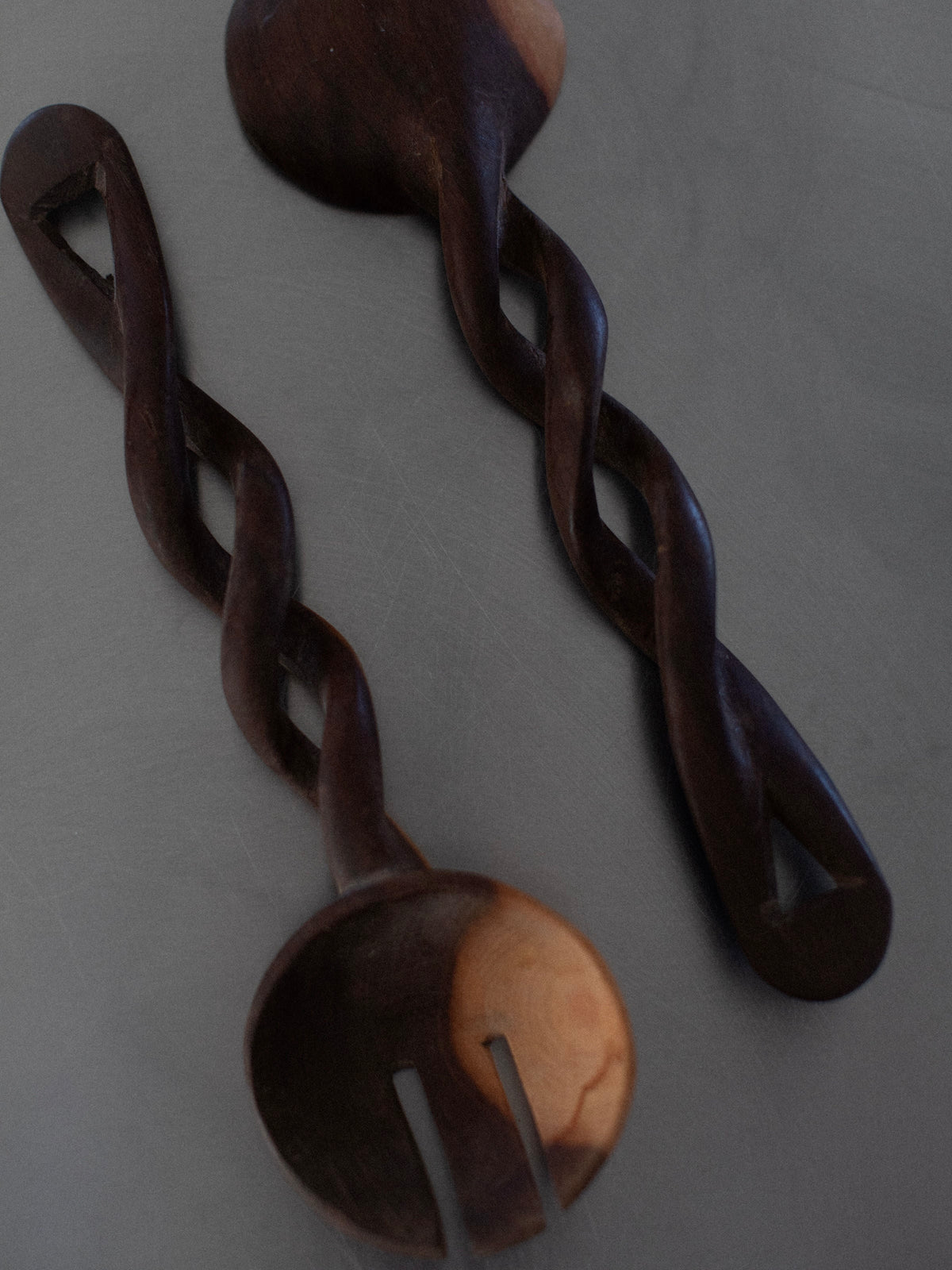 The Out For Lunch Twisted Serving Spoons, a pair of dark wooden utensils featuring a spoon and fork with intricately twisted handles, lie on a gray surface. These handcrafted culinary tools showcase a natural grain that varies in color, adding a rustic feel to their elegant design.