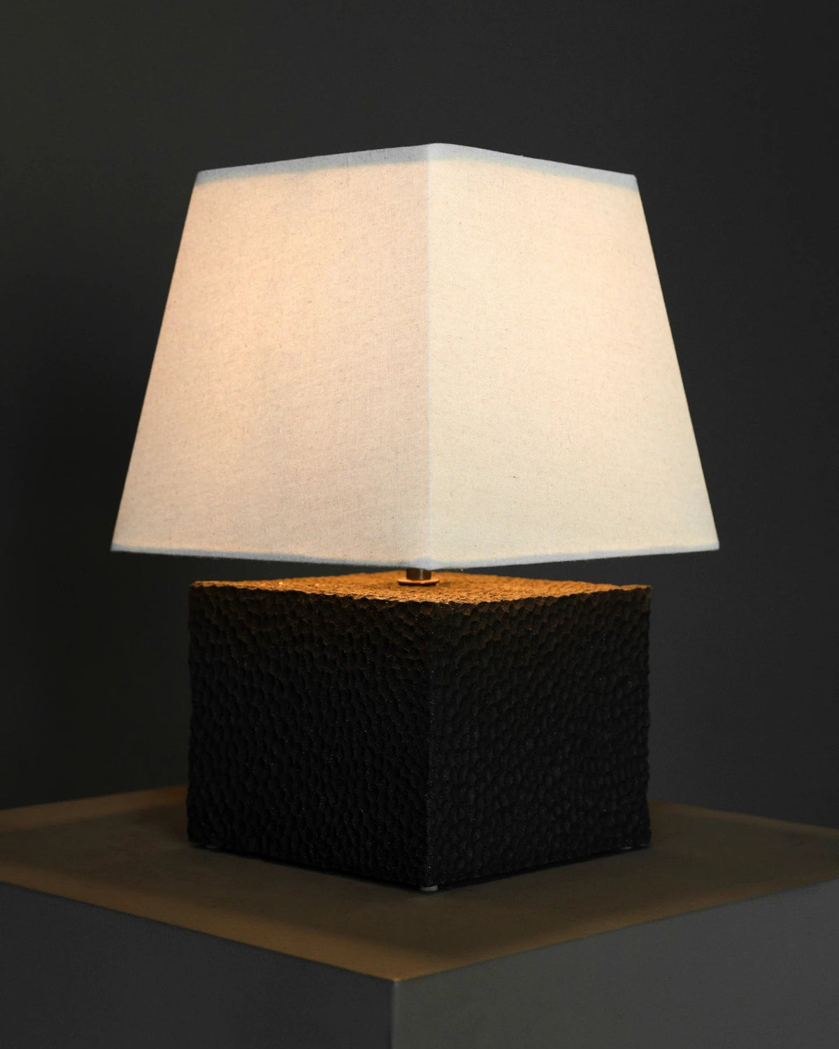 The Akune Ceramic Lamp - Number Five by Studio OSKLO, featuring a white square lampshade and a textured black square base inspired by post-Bauhaus brutalism, sits illuminated on a dark surface against a dark gray background.