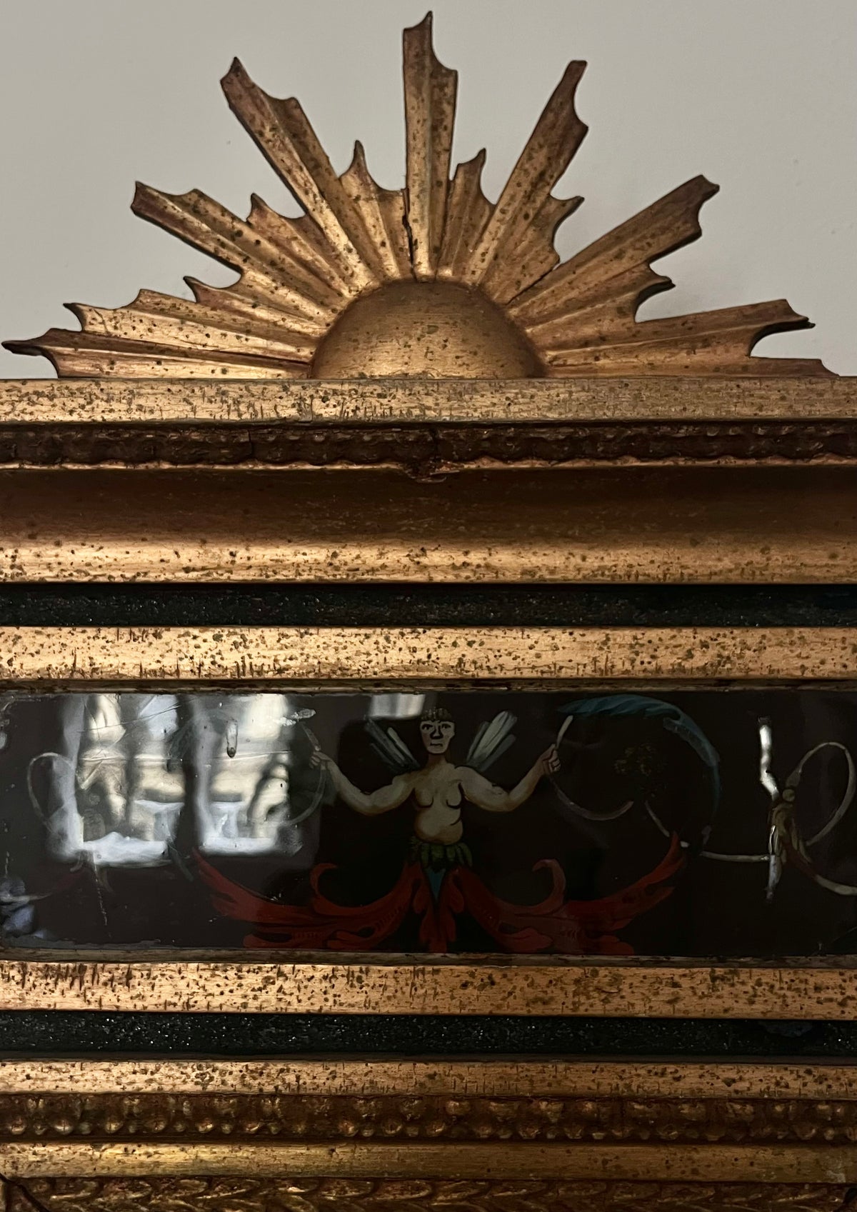 The Médecine Antique Gilded Mirror showcases an ornate, sunburst-topped frame and a painted panel below featuring a winged figure with outstretched arms, set amid dark and red hues.