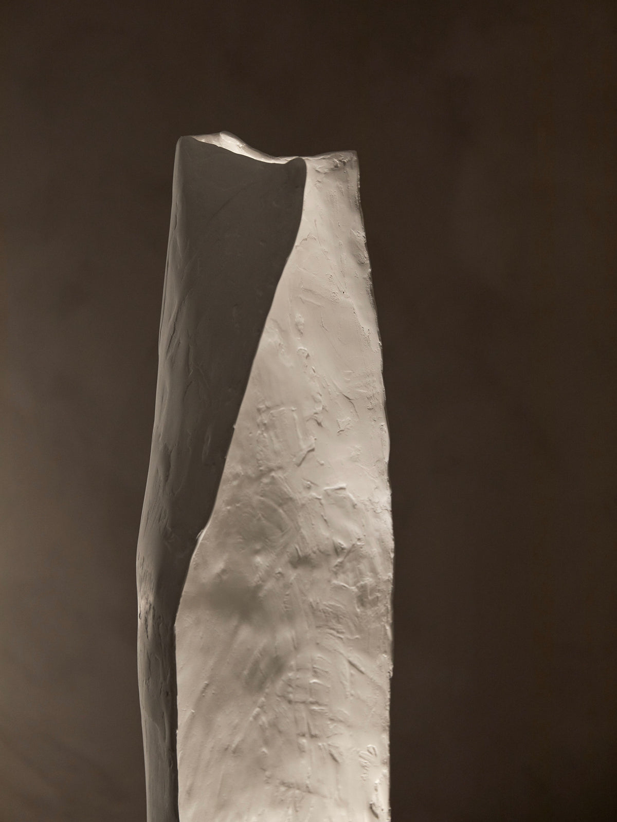 The Alcove Floor Lamp by Studio Tristan Kallas features a tall, angular design against a dark background. Its textured surfaces and sharp edges create contrasting shadows, exuding a contemporary, minimalist aesthetic with subtly intertwined organic shapes.