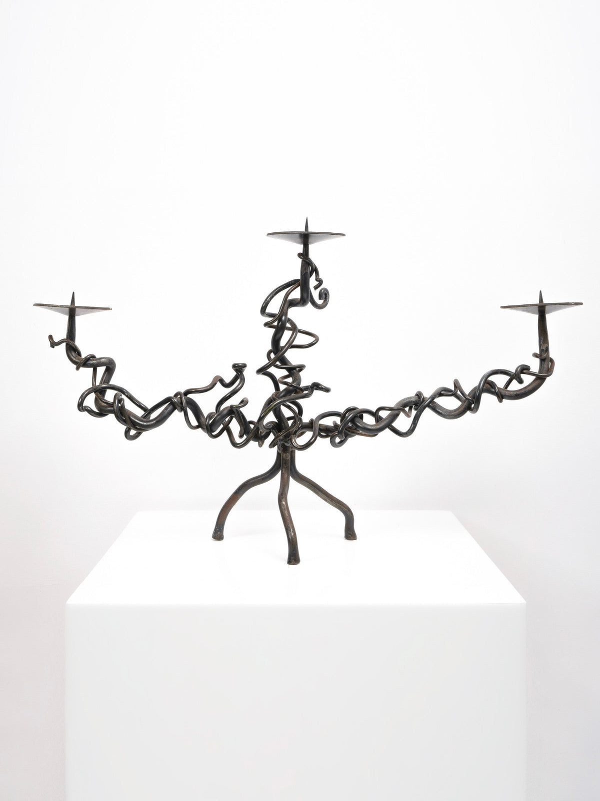 The Tom Bogle Brutalist Iron Candelabra, featuring three vine-like curling arms, is displayed on a white pedestal against a plain white background.
