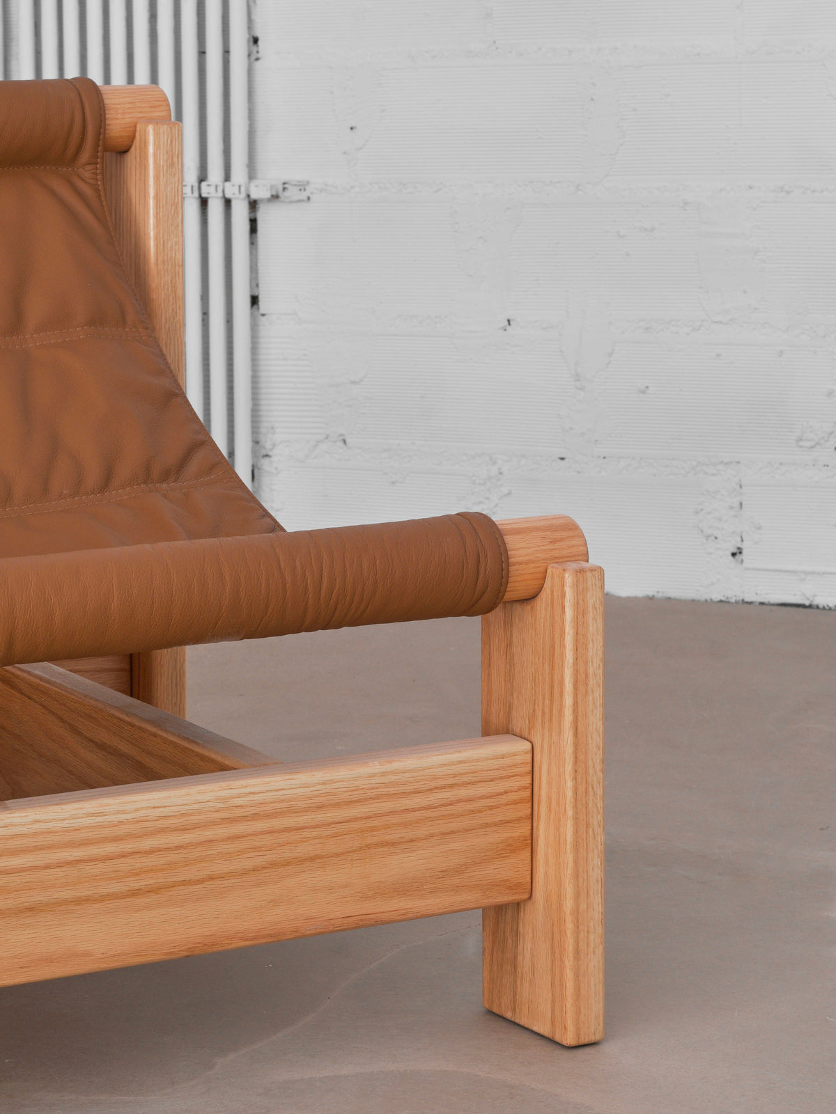 Close-up of malva's "Lounge Up" Lounge Chair, featuring a light oak frame and tan leather upholstery for adaptive comfort. Shown against a light wall and concrete floor.