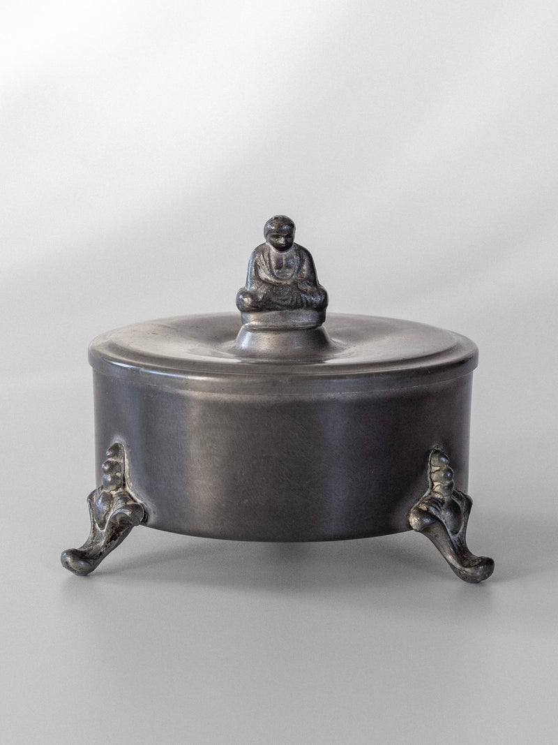A round Art Deco pewter box with a lid showcases a small seated Buddha figure on top. Designed by Spigel, it is supported by three ornate legs, providing an elevated look against a neutral gray background.