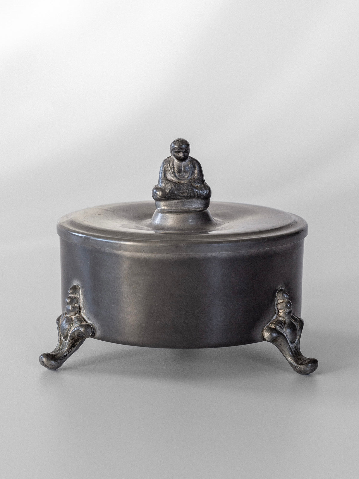 A round Art Deco pewter box with a lid showcases a small seated Buddha figure on top. Designed by Spigel, it is supported by three ornate legs, providing an elevated look against a neutral gray background.
