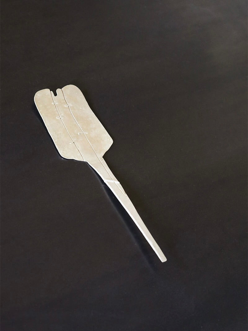 A Silver Plated Cake Server from Rooms Studio lies on a dark surface. The cake server features a sleek, elongated shape with a slight curve at the broader end and a handle that tapers to a narrow point, resembling an elegant shoehorn with a minimalist design you might find in Tbilisi, Georgia.
