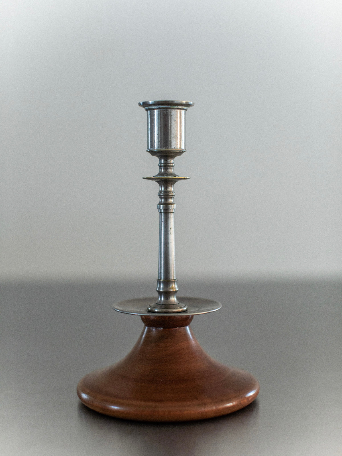The Out For Lunch Steel & Wood Candleholders feature a metallic design on a wide, round wooden base, exuding rustic charm. Elegantly positioned on a smooth gray surface, they embody the timeless appeal of classic candleholders against a plain background.